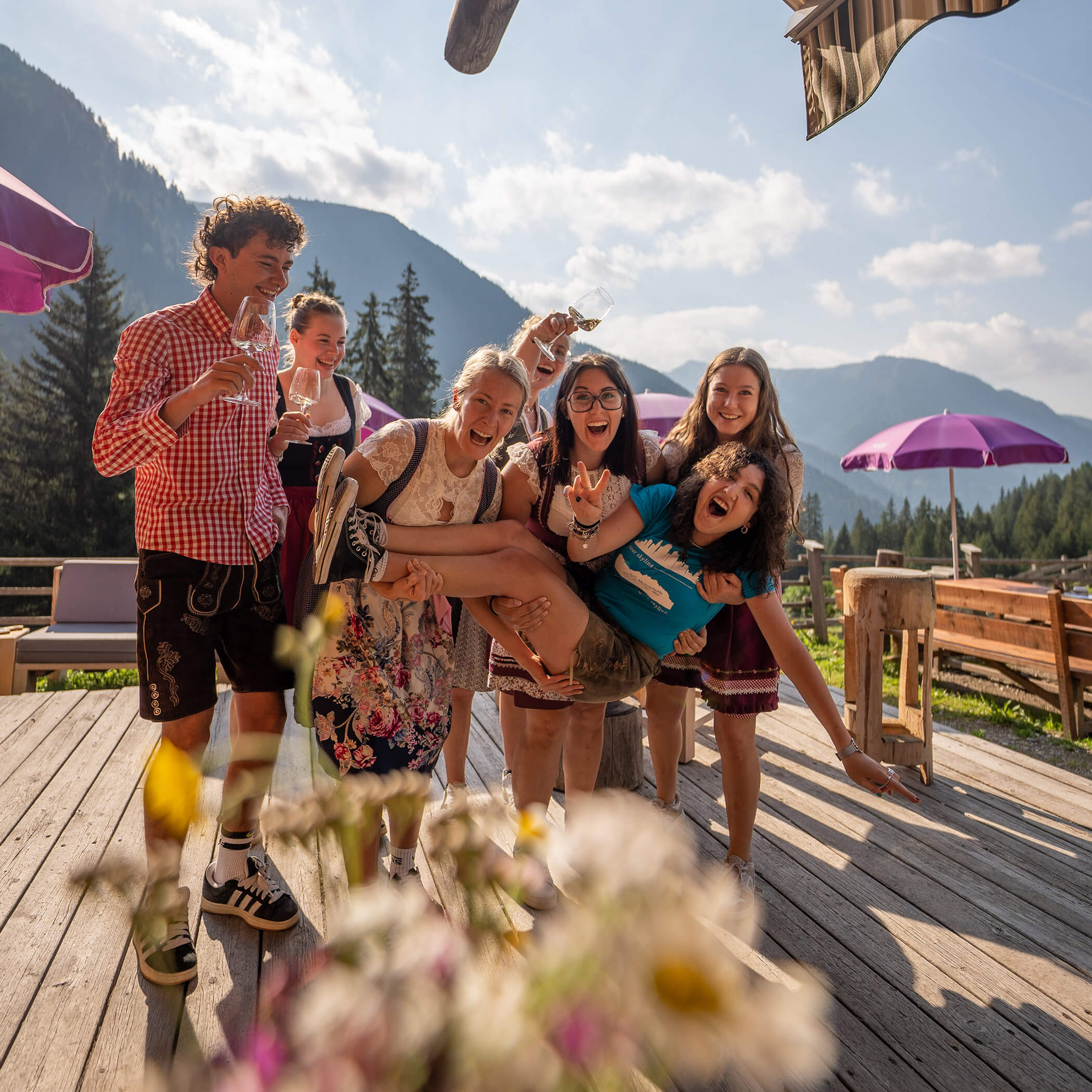 Eine Gruppe lächelnder Freunde im Freien hebt eine Person hoch, mit Bergen und lila Regenschirmen im Hintergrund. - Hotel La Casies Mountain Living