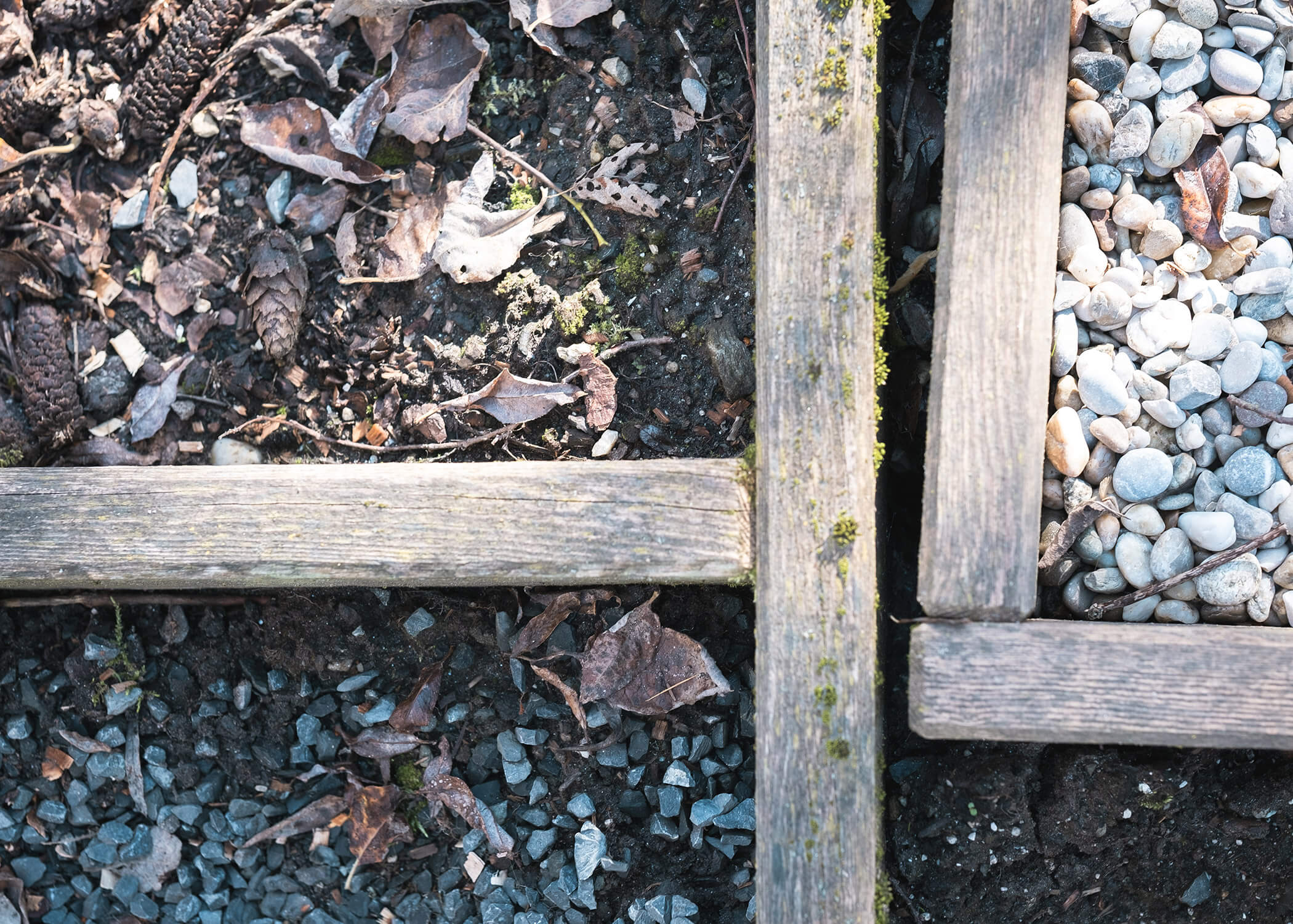 Nahaufnahme einer hölzernen Garteneinfassung, die Erde, trockenes Laub, Kies und kleine weiße Steine in Abschnitte unterteilt. - Hotel La Casies Mountain Living