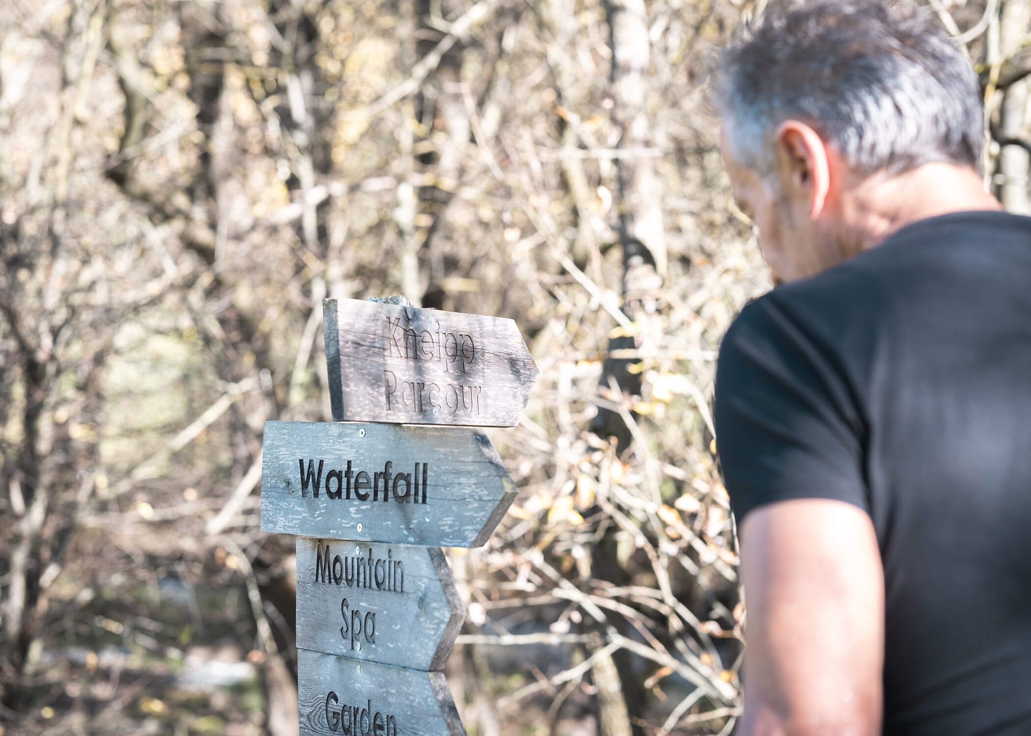 Ein Mann schaut auf einen hölzernen Wegweiser im Wald, der auf einen Wasserfall, ein Bergbad und einen Garten hinweist. - Hotel La Casies Mountain Living