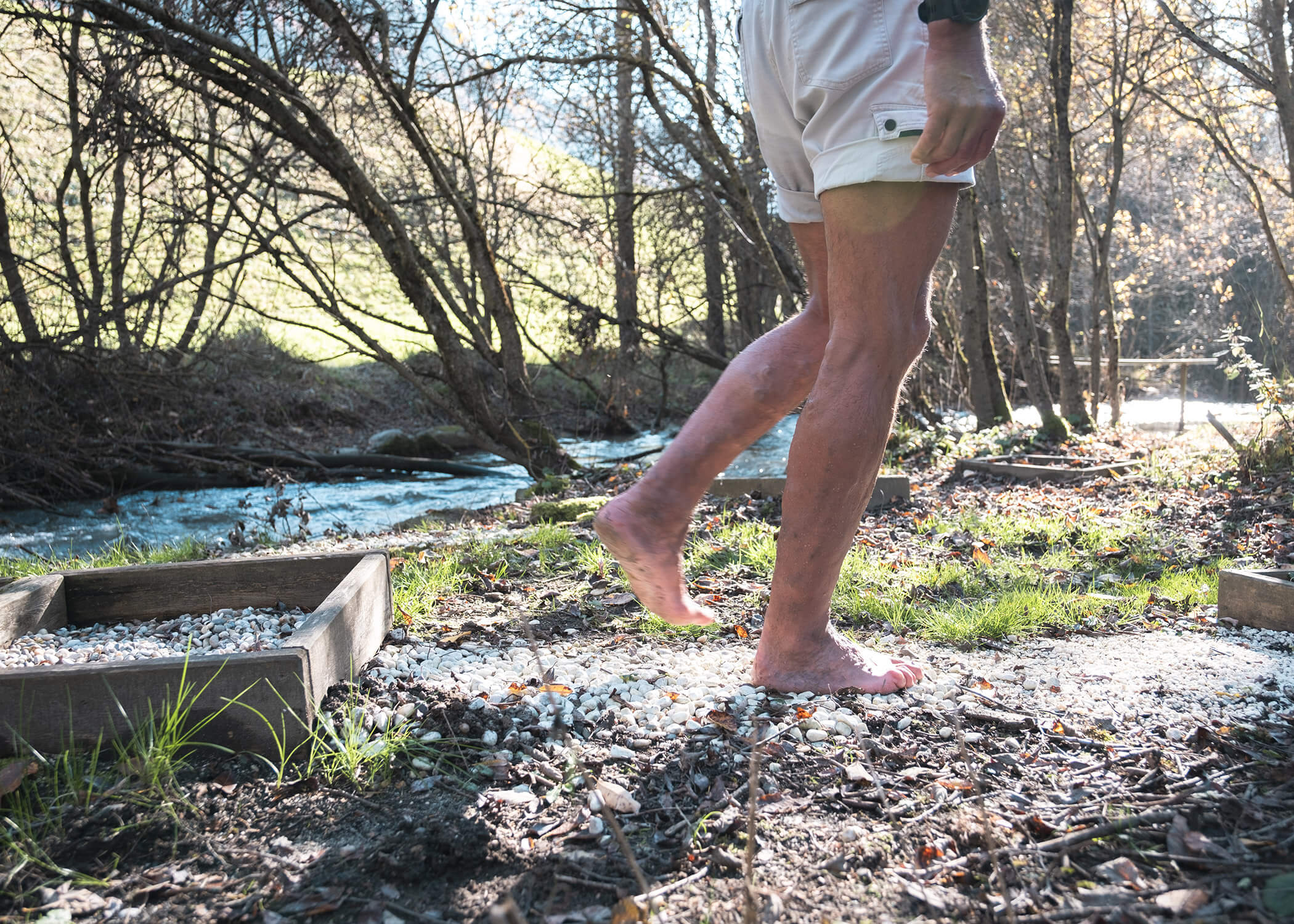 Eine Person geht barfuß auf einem Kiesweg neben einem Bach in einem sonnenbeschienenen, bewaldeten Gebiet. - Hotel La Casies Mountain Living