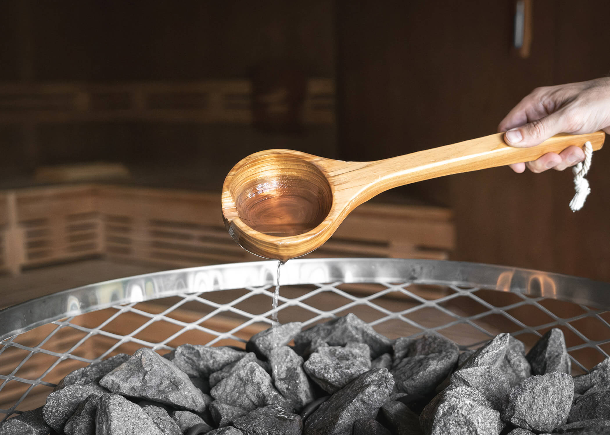 Mit einer Holzkelle wird Wasser auf heiße Saunasteine über einem Metallrost gegossen, wodurch Dampf entsteht. - Hotel La Casies Mountain Living