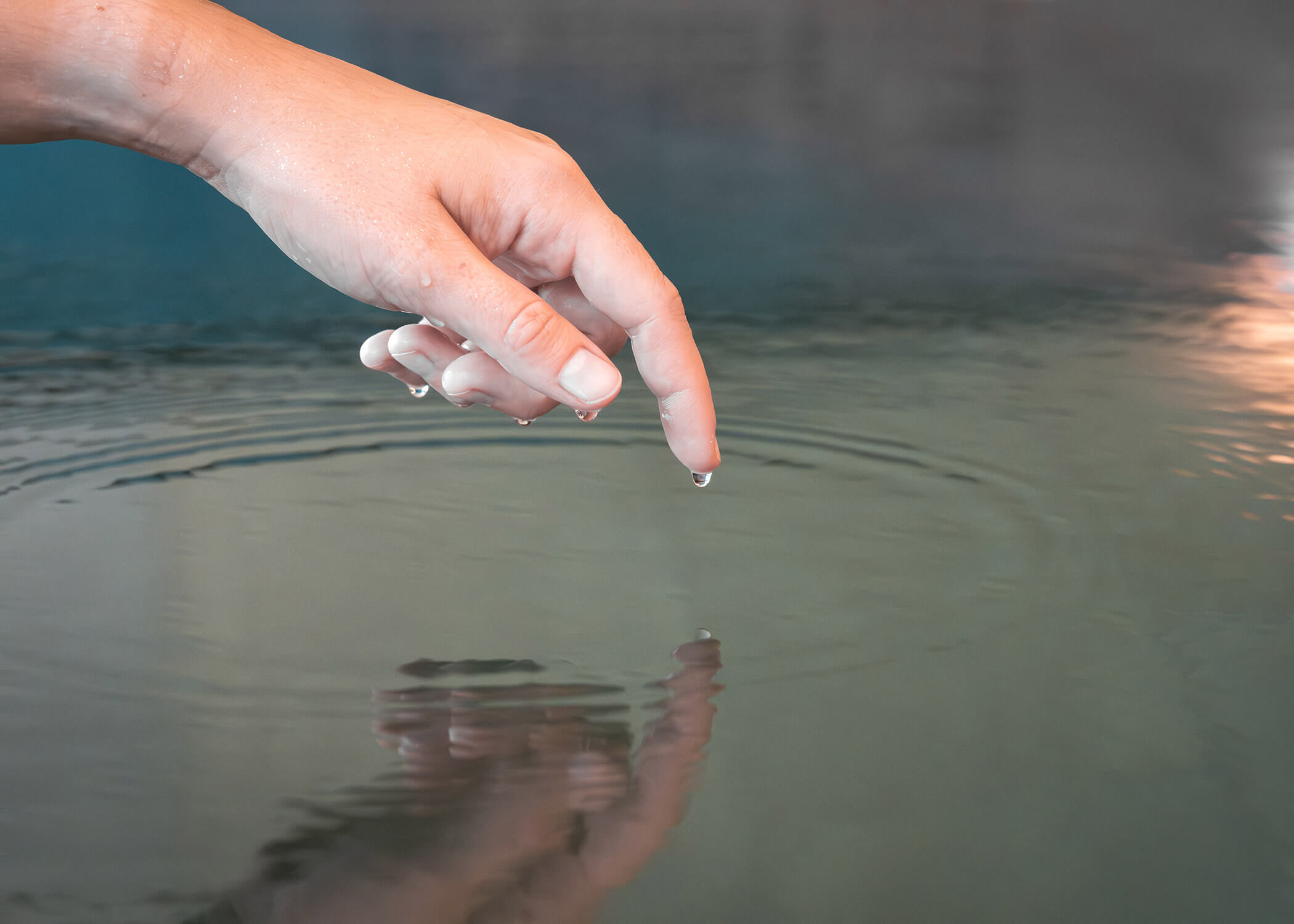 Eine Hand berührt sanft das ruhige Wasser, wobei ein Tropfen von der Fingerspitze fällt und sich kräuselt. - Hotel La Casies Mountain Living