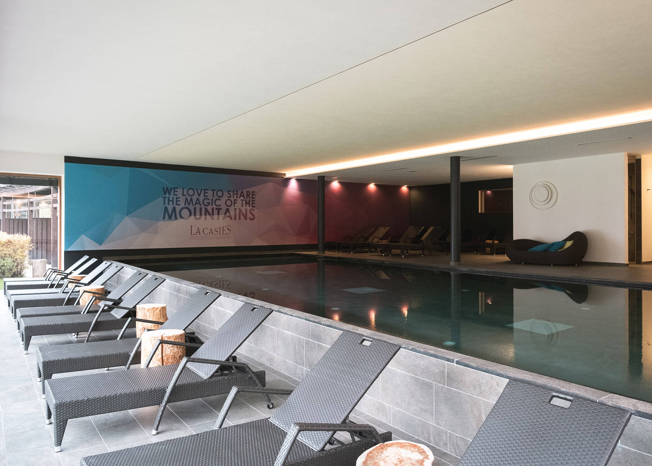 Innenpoolbereich mit Liegestühlen, Holzhockern und einem Wandgemälde über den Zauber der Berge. - Hotel La Casies Mountain Living