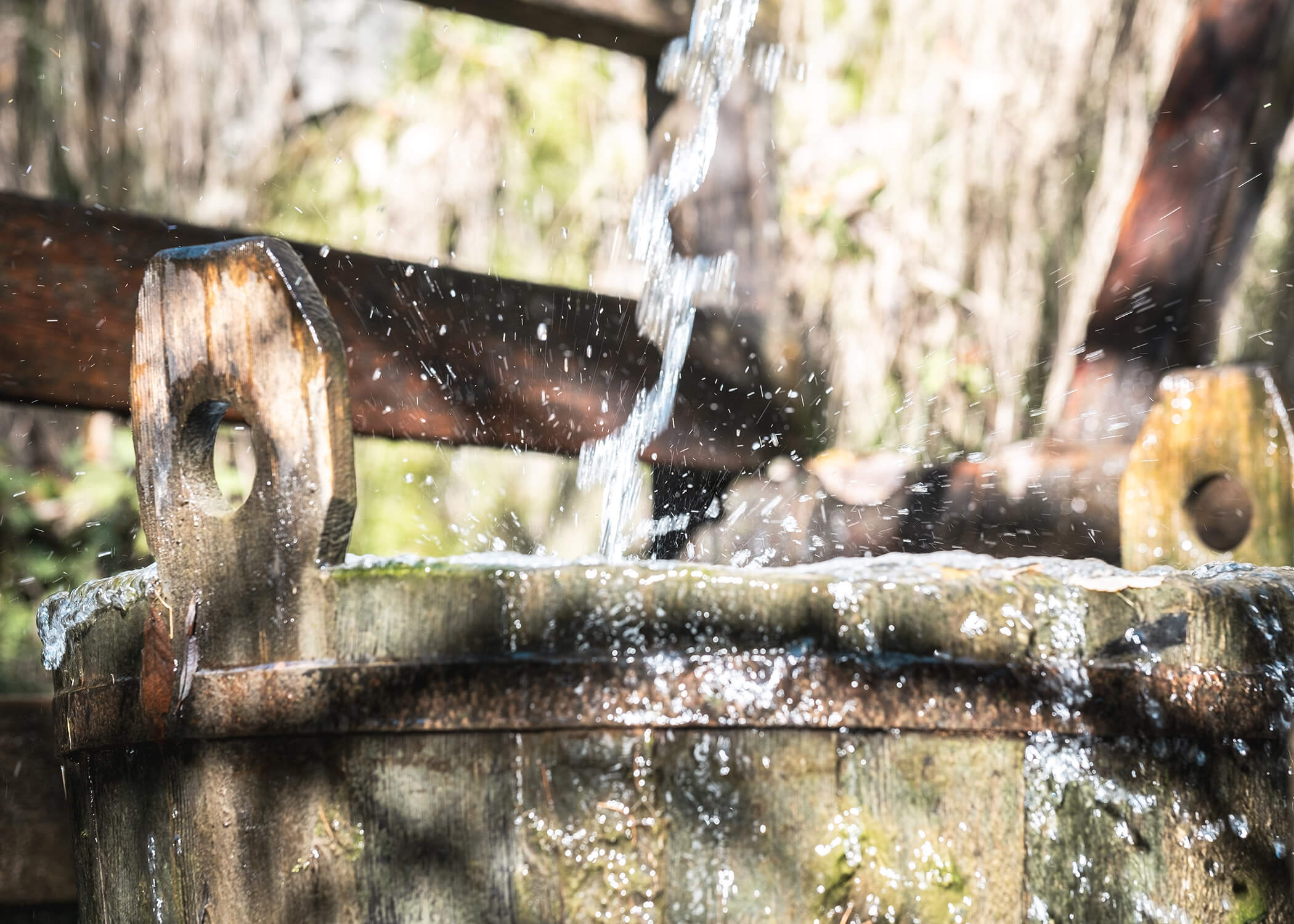Wasser, das im Freien in einen Holzeimer spritzt, wobei das Sonnenlicht das fließende Wasser und die Holzstruktur hervorhebt. - Hotel La Casies Mountain Living