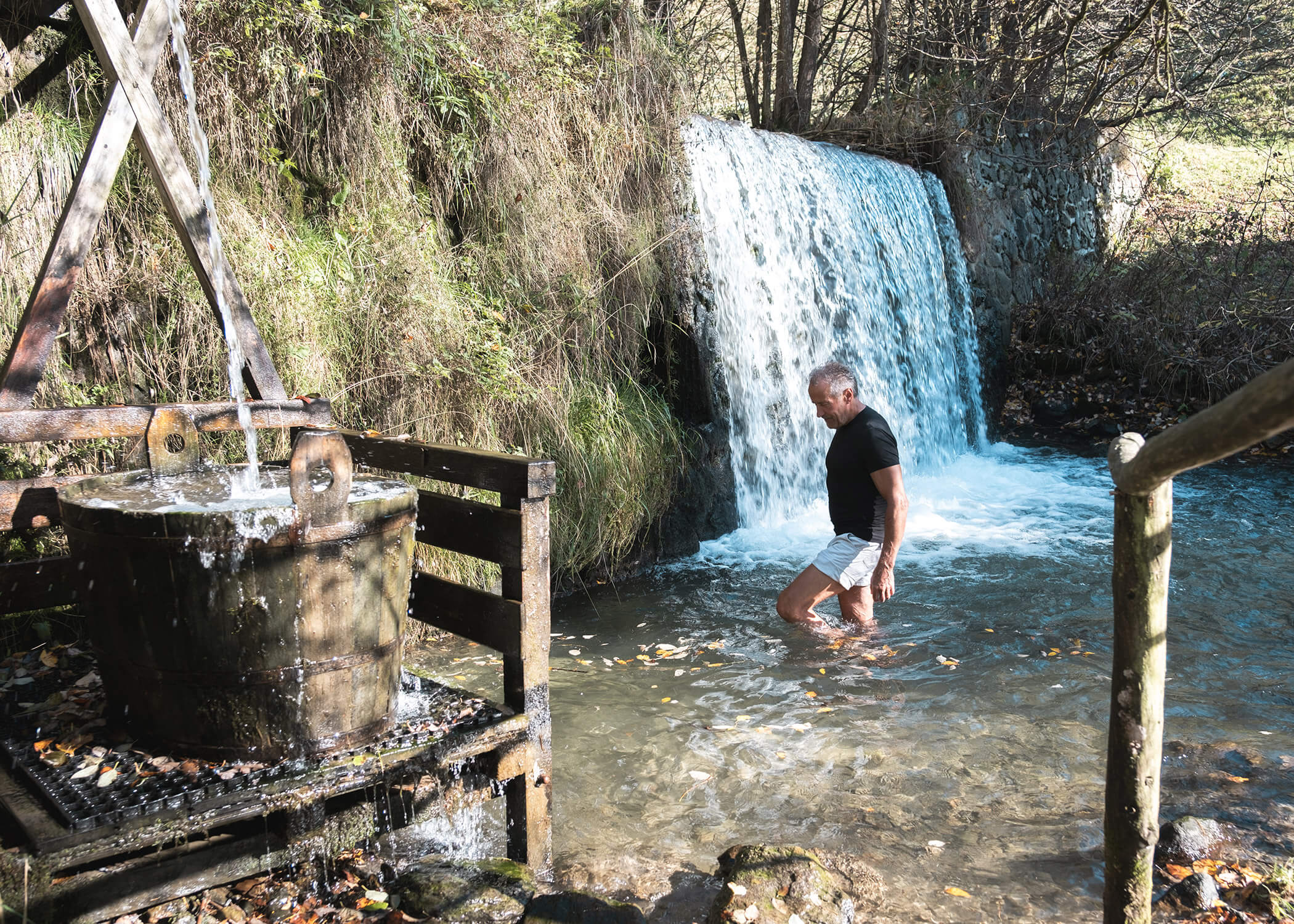 Ein Mann steht in einem seichten Bach in der Nähe eines kleinen Wasserfalls und einer hölzernen Wassermühle in einer natürlichen Umgebung. - Hotel La Casies Mountain Living