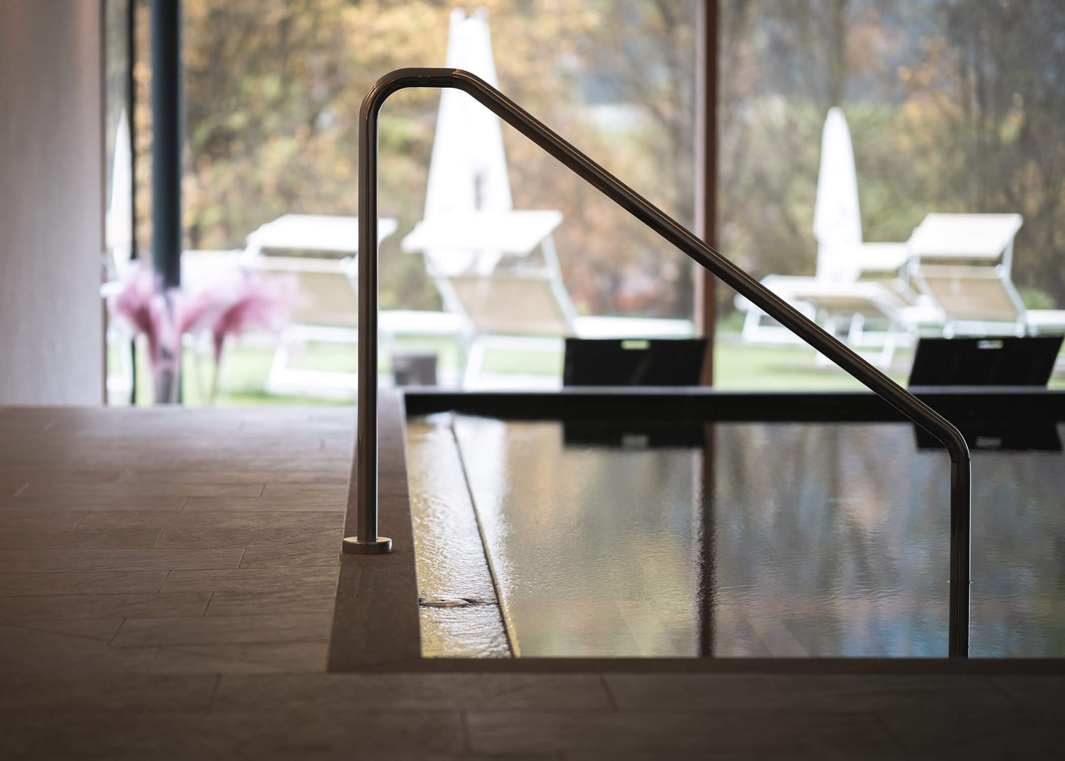 Hallenbad mit Metallgeländer, mit Blick auf einen Garten mit Liegestühlen und Sonnenschirmen im Freien. - Hotel La Casies Mountain Living