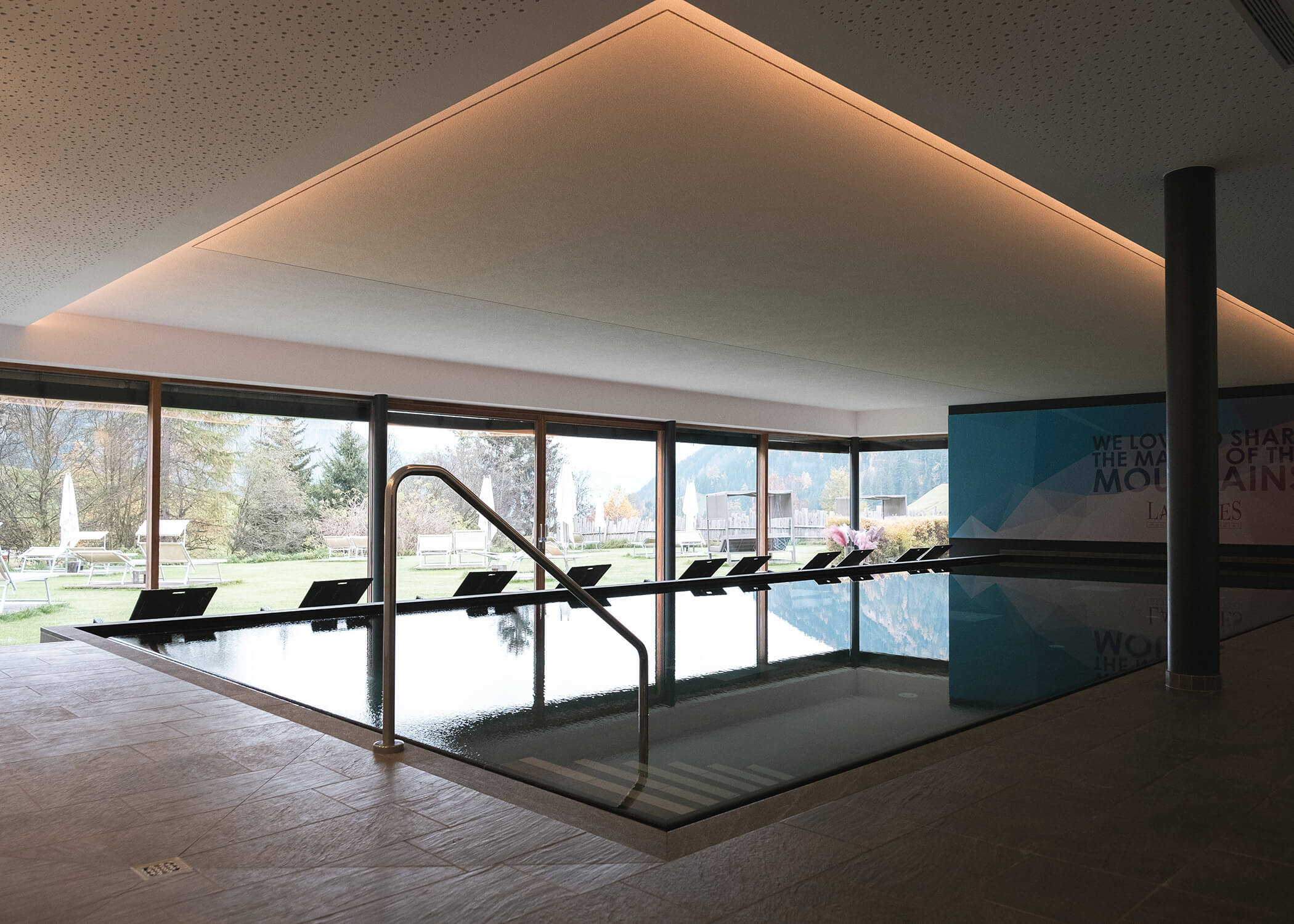 Innenpool mit Liegestühlen an großen Fenstern mit Blick auf einen grasbewachsenen Außenbereich und Bäume. - Hotel La Casies Mountain Living