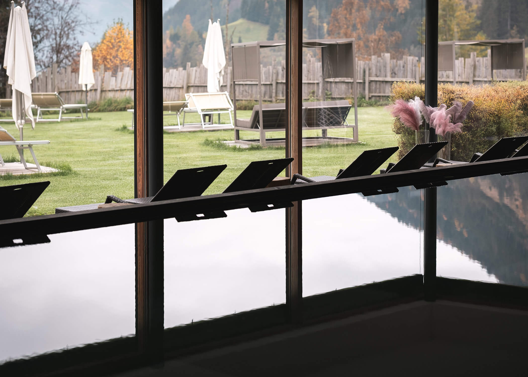 Die Sessel am Fenster bieten einen Blick auf einen grasbewachsenen Außenbereich mit Sonnenschirmen und Holzzäunen. - Hotel La Casies Mountain Living