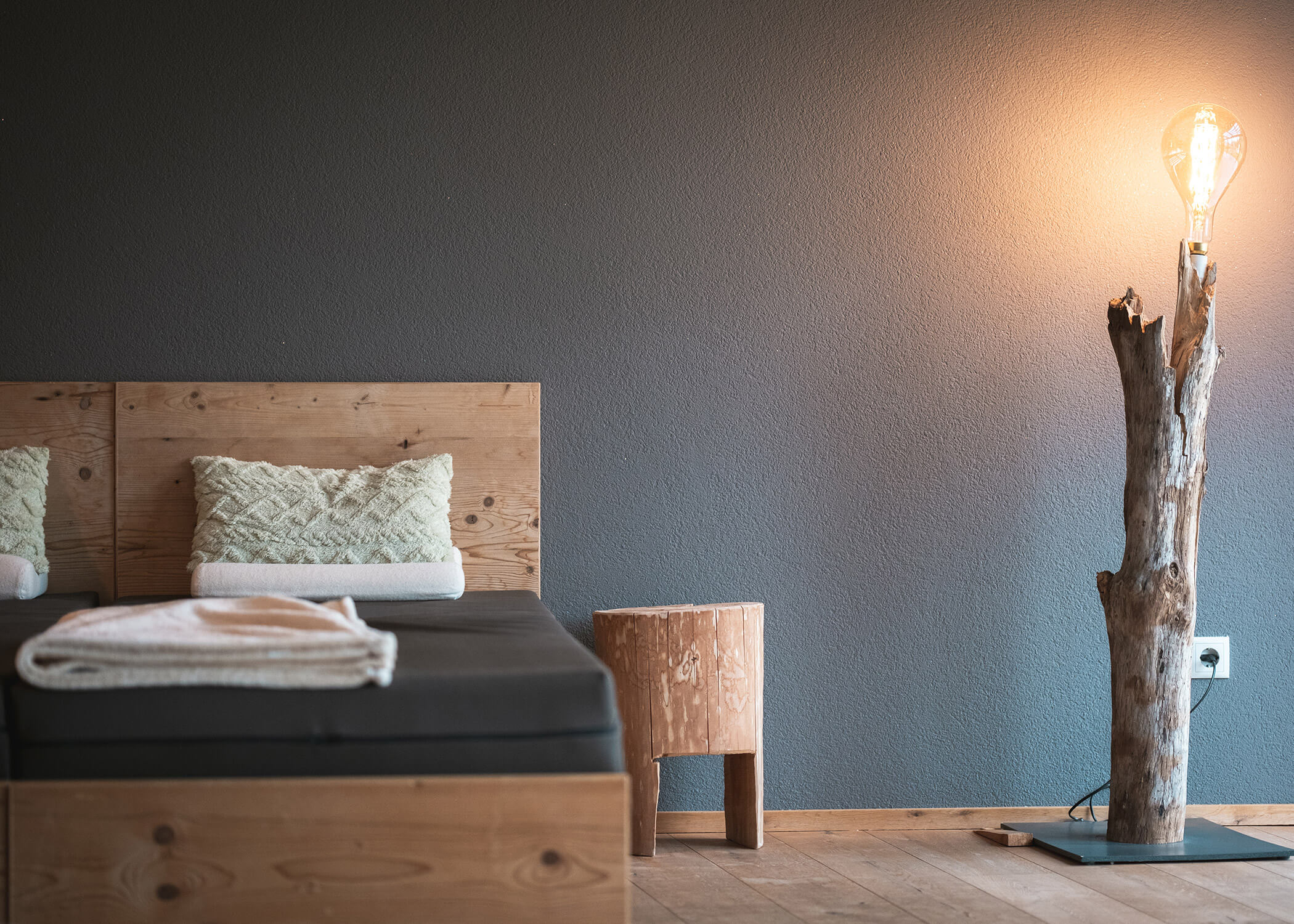 Minimalistisches Schlafzimmer mit Holzmöbeln, einem Bett, einem kleinen Hocker und einer rustikalen Stehlampe in Form eines Baumstamms. - Hotel La Casies Mountain Living