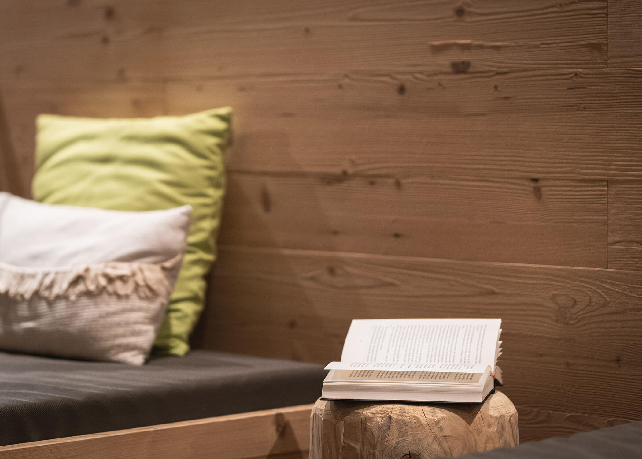 Eine gemütliche Sitzecke mit Kissen und einem aufgeschlagenen Buch, das auf einem Holzhocker neben einer holzgetäfelten Wand liegt. - Hotel La Casies Mountain Living