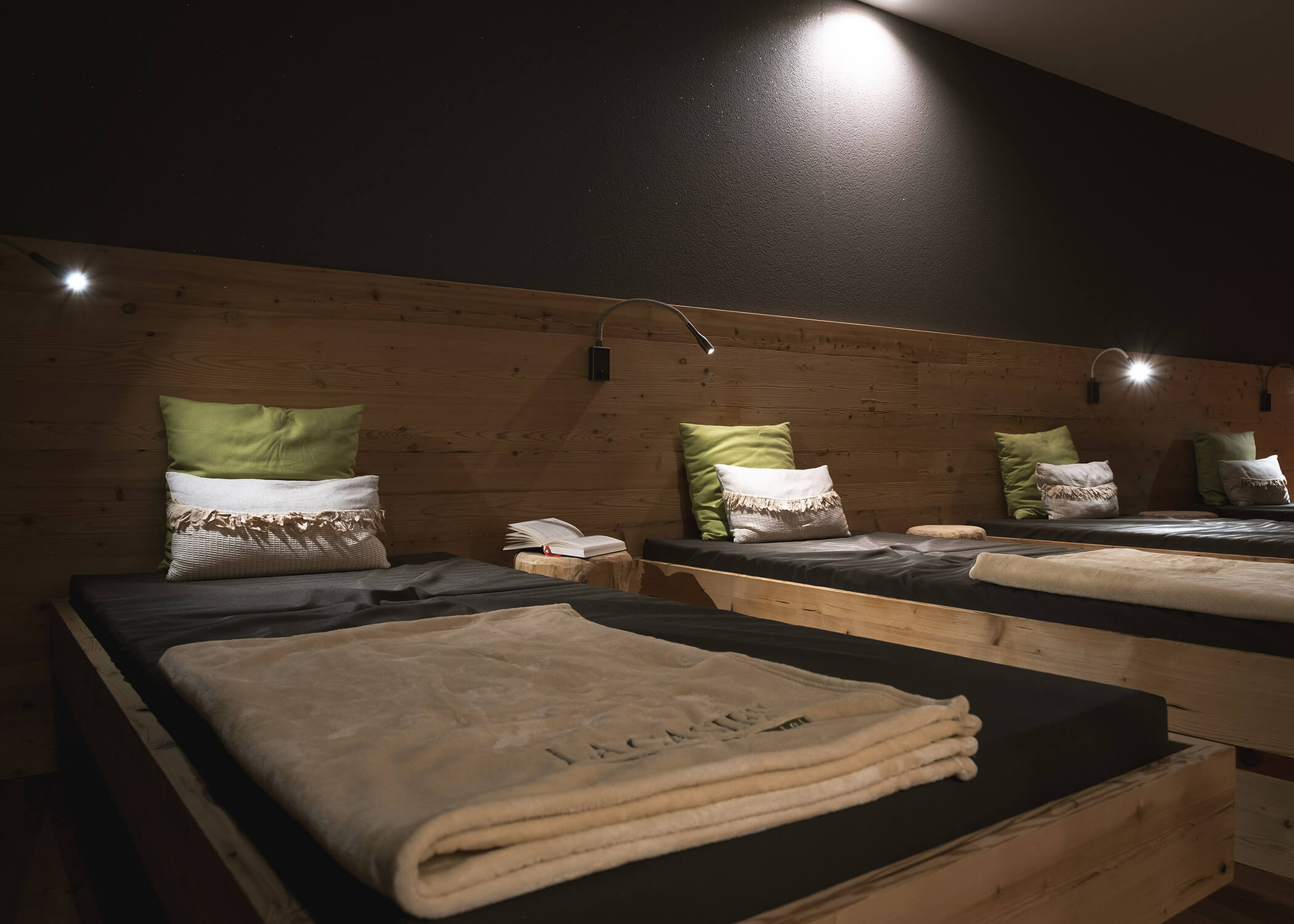 Eine Reihe gemütlicher Betten mit grünen Kissen und Decken in einem schummrigen, hölzernen Ruheraum. - Hotel La Casies Mountain Living