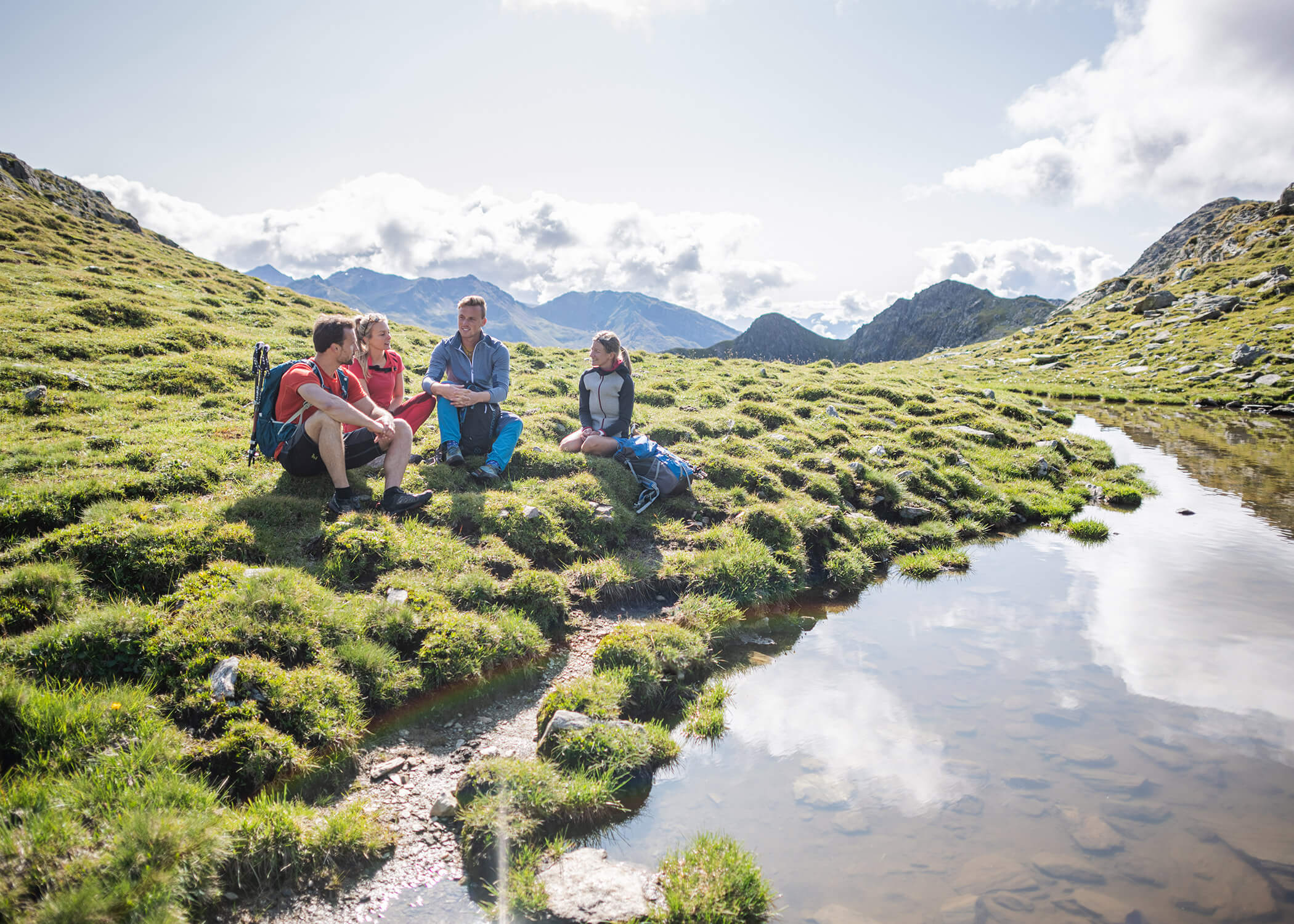 Vier Personen mit Rucksäcken sitzen im Gras an einem Teich in einer bergigen Landschaft unter einem teilweise bewölkten Himmel. - Hotel La Casies Mountain Living