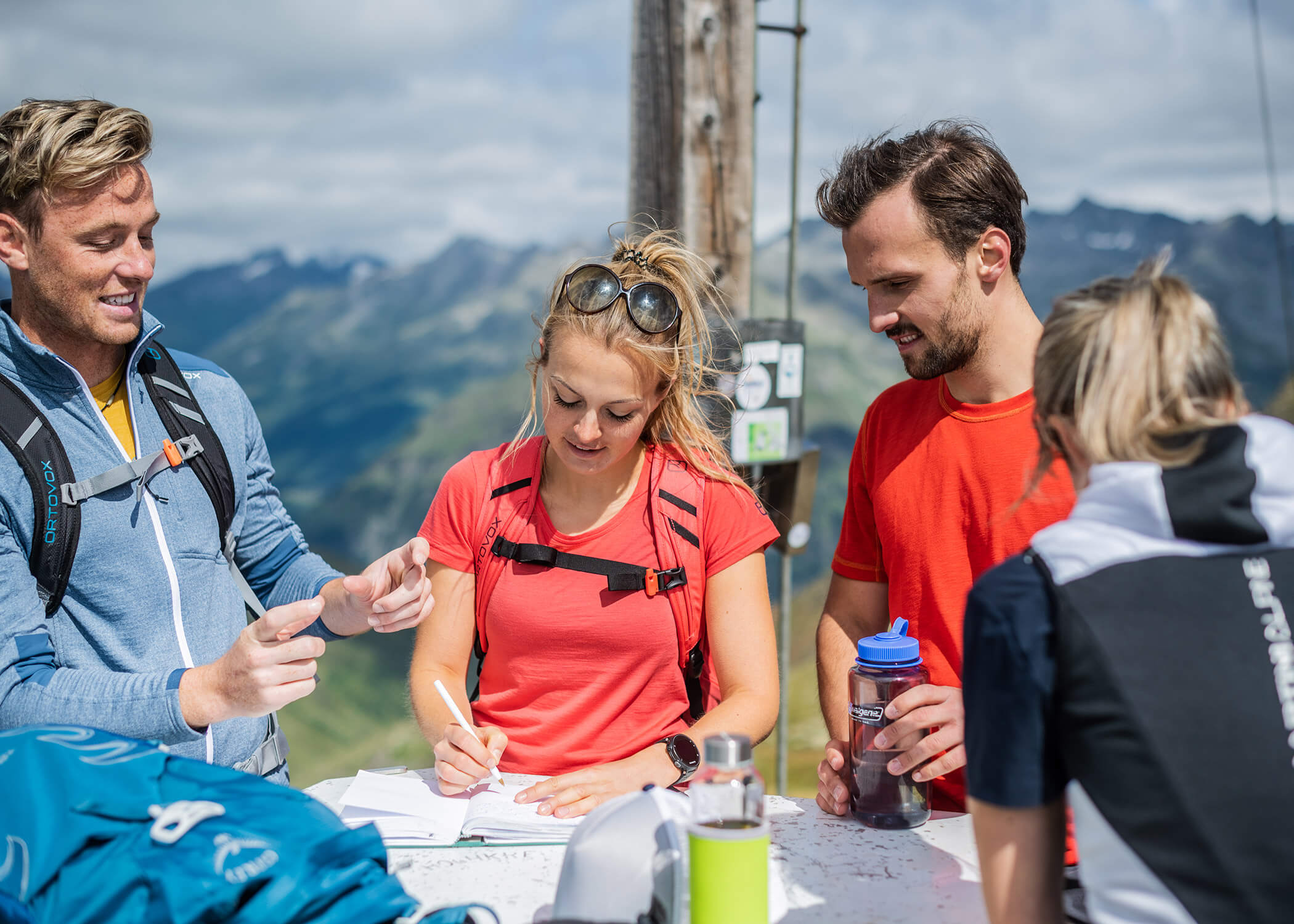 Vier Wanderer lächeln und schreiben an einem sonnigen Tag auf einem Berggipfel in ein Notizbuch. - Hotel La Casies Mountain Living