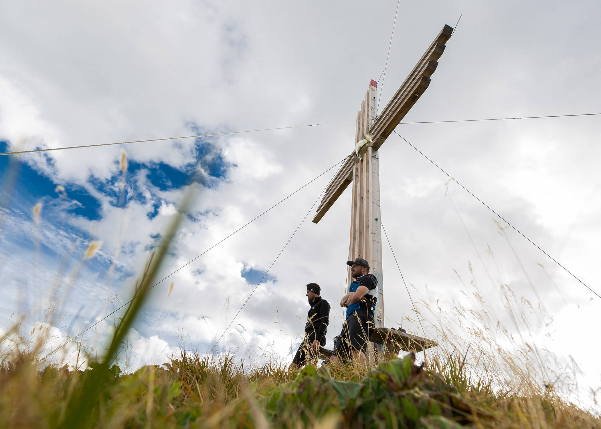 Zwei Personen stehen bei einem großen Holzkreuz auf einem grasbewachsenen Hügel unter einem teilweise bewölkten Himmel. - Hotel La Casies Mountain Living