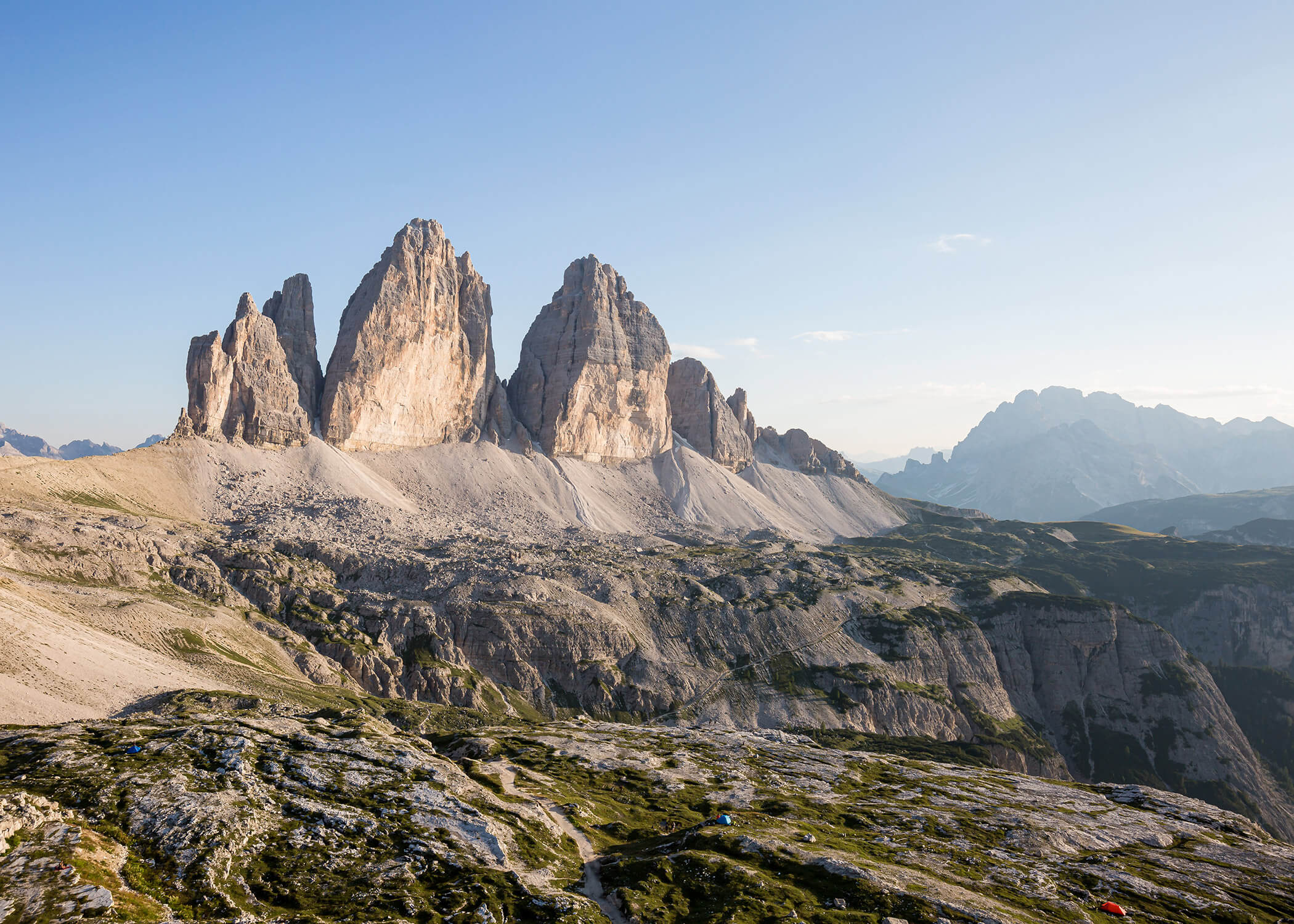 Drei felsige Berggipfel erheben sich über eine zerklüftete, sonnenbeschienene Landschaft mit klarem Himmel im Hintergrund. - Hotel La Casies Mountain Living