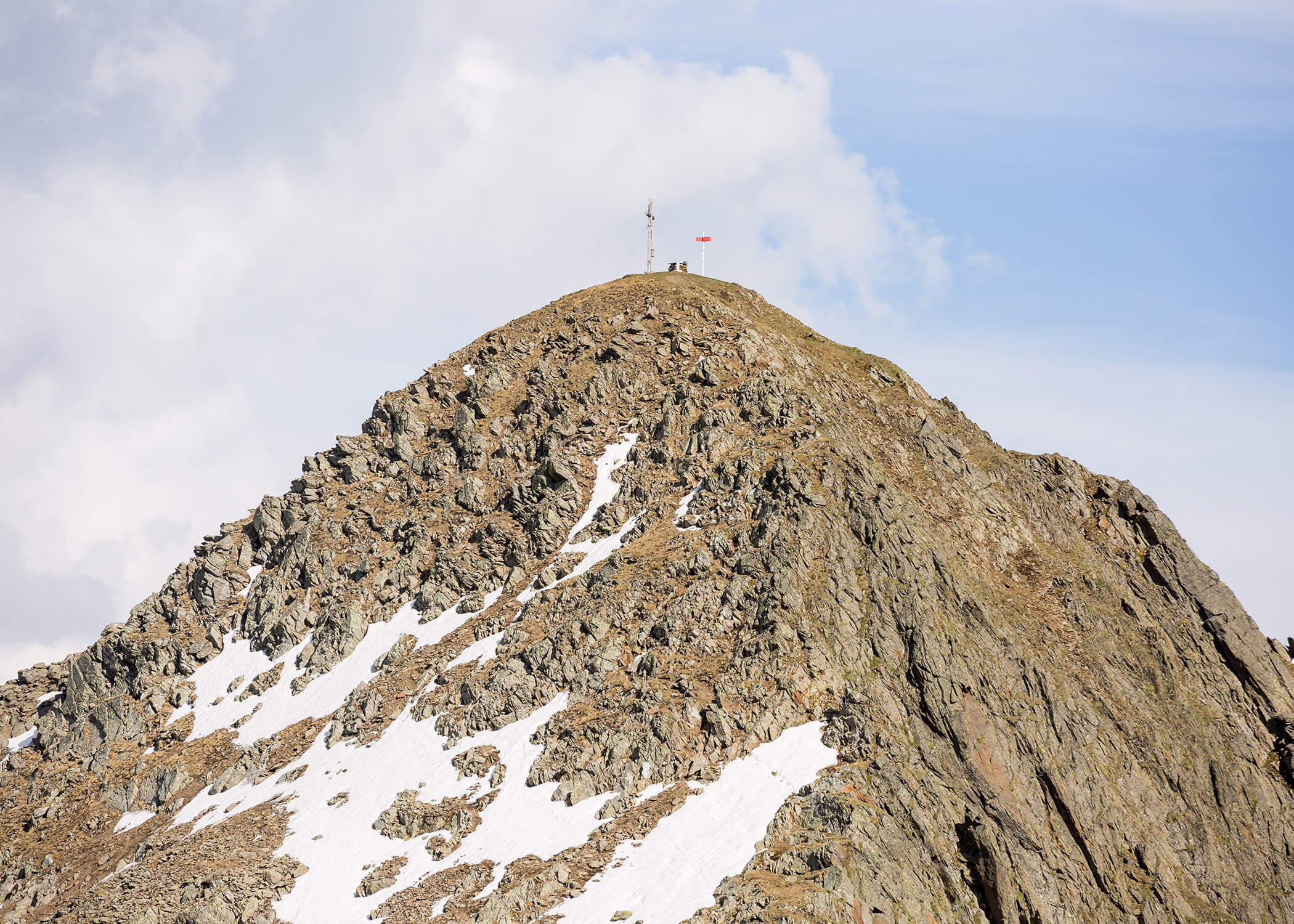 Felsengipfel mit Schneeflecken, einem Gipfelkreuz und einer kleinen Fahne unter einem teilweise bewölkten Himmel. - Hotel La Casies Mountain Living