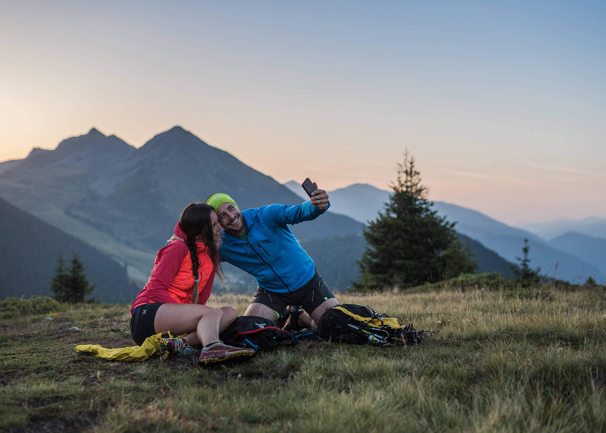 Zwei Wanderer sitzen im Gras und machen ein Selfie mit Bergen und Bäumen im Hintergrund bei Sonnenuntergang. - Hotel La Casies Mountain Living