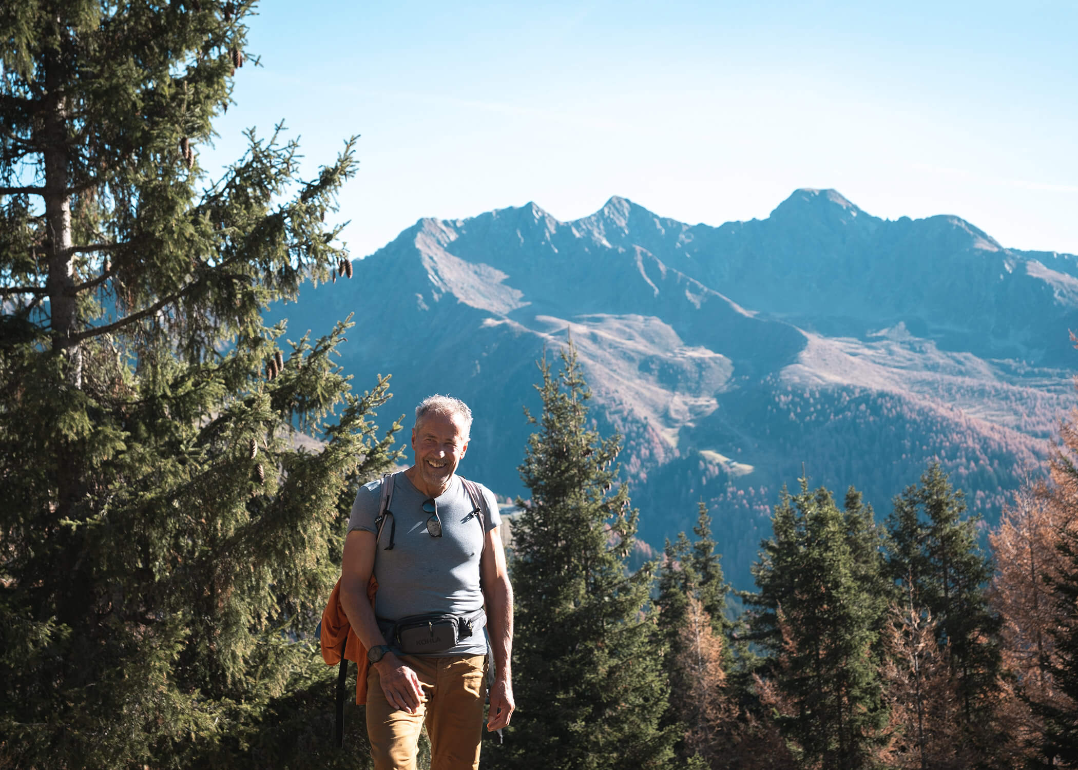 Lächelnder Mann beim Wandern in einem bewaldeten Berggebiet mit hohen Bäumen und fernen felsigen Gipfeln im Sonnenlicht. - Hotel La Casies Mountain Living
