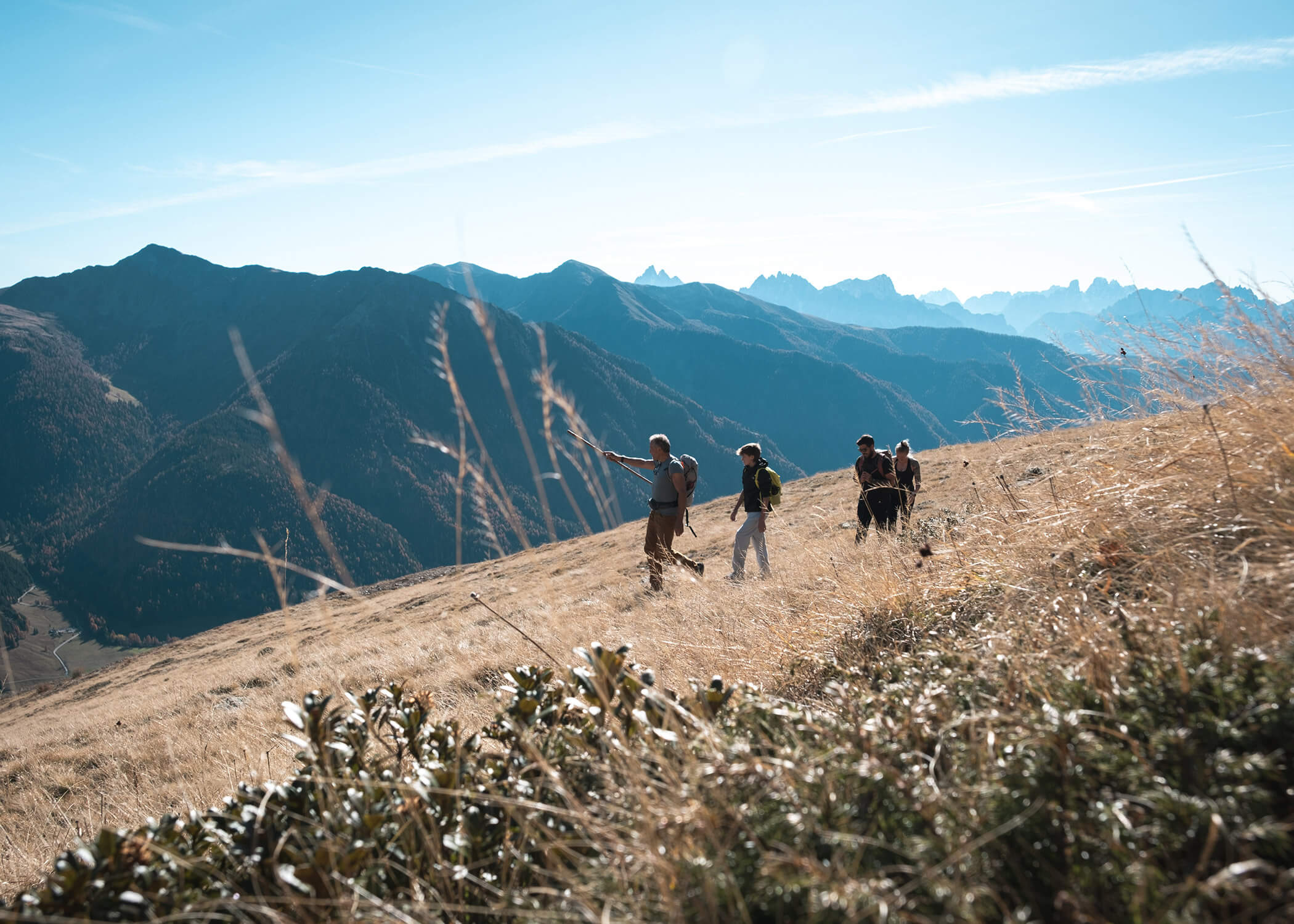 Vier Wanderer wandern an einem grasbewachsenen Berghang mit weit entfernten Gipfeln unter einem klaren blauen Himmel. - Hotel La Casies Mountain Living