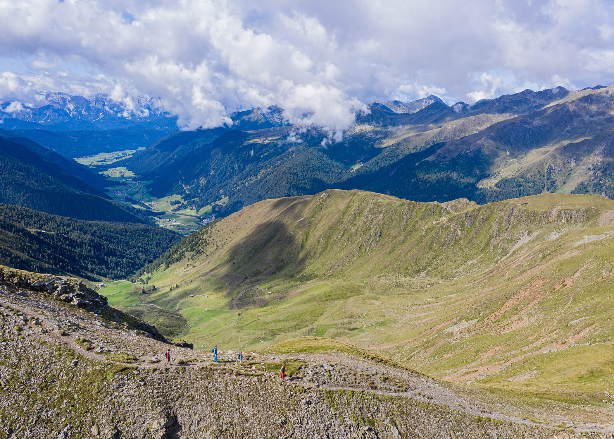 Wanderer wandern auf einem Bergpfad mit grünen Tälern und fernen Gipfeln unter einem teilweise bewölkten Himmel. - Hotel La Casies Mountain Living