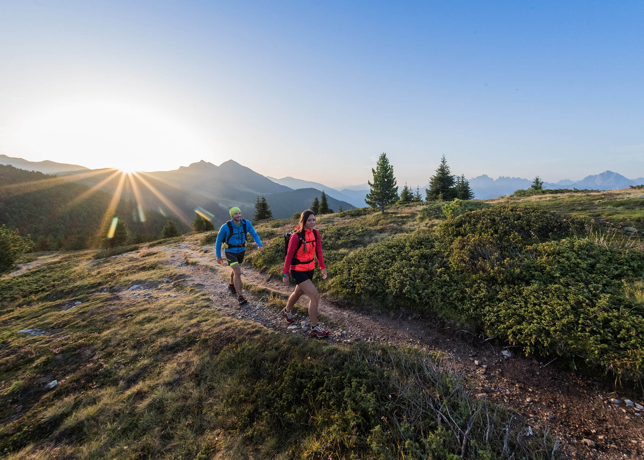 Zwei Wanderer gehen bei Sonnenaufgang einen Bergpfad entlang, umgeben von Grün und fernen Gipfeln. - Hotel La Casies Mountain Living