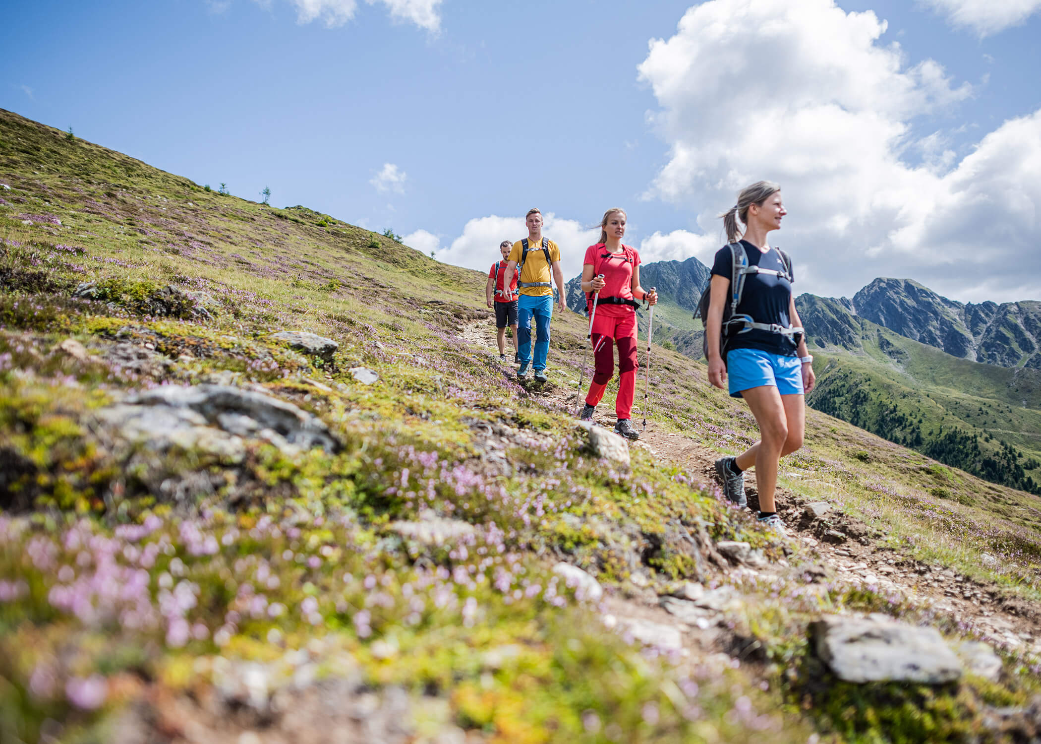 Vier Personen wandern auf einem grasbewachsenen Bergpfad unter einem blauen Himmel mit Wolken und fernen Felsgipfeln. - Hotel La Casies Mountain Living