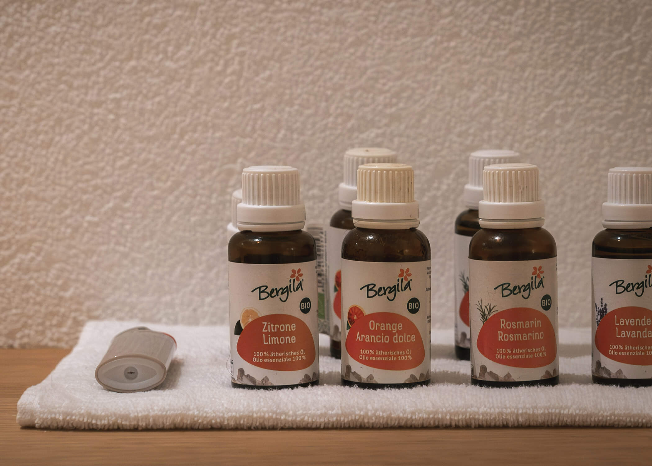 Sechs kleine Flaschen ätherischer Öle mit italienischen und deutschen Etiketten auf einem weißen Handtuch vor einer strukturierten Wand. - Hotel La Casies Mountain Living