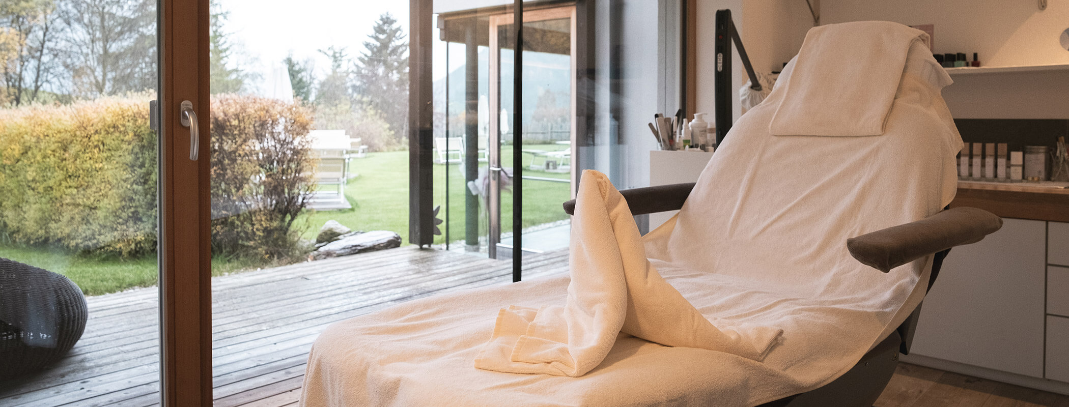 Ein mit weißen Handtüchern bedeckter Spa-Behandlungsstuhl neben großen Fenstern mit Blick auf einen Garten. - Hotel La Casies Mountain Living