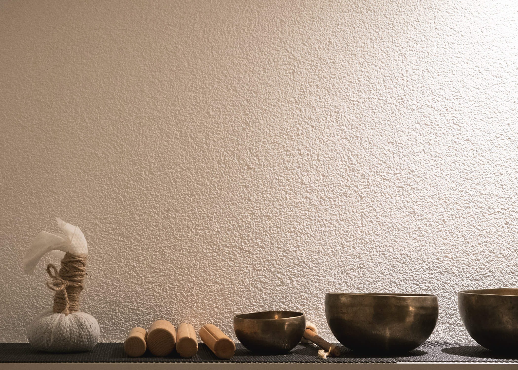 Drei Klangschalen, Massagestäbchen und eine Kräuterkompresse auf einer Matte vor einer strukturierten beigen Wand. - Hotel La Casies Mountain Living