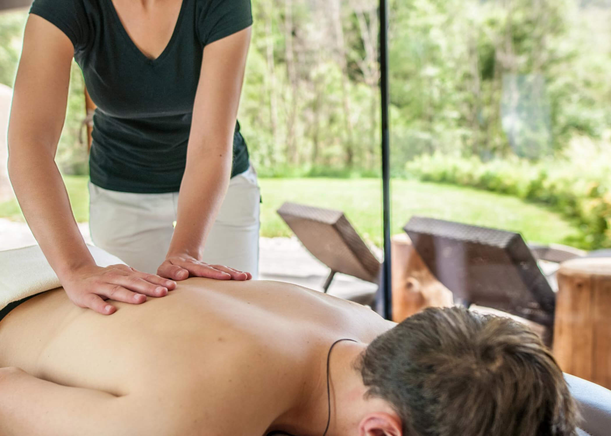Eine Person erhält eine Rückenmassage in einem hellen, entspannenden Raum mit großen Fenstern und Blick ins Grüne. - Hotel La Casies Mountain Living