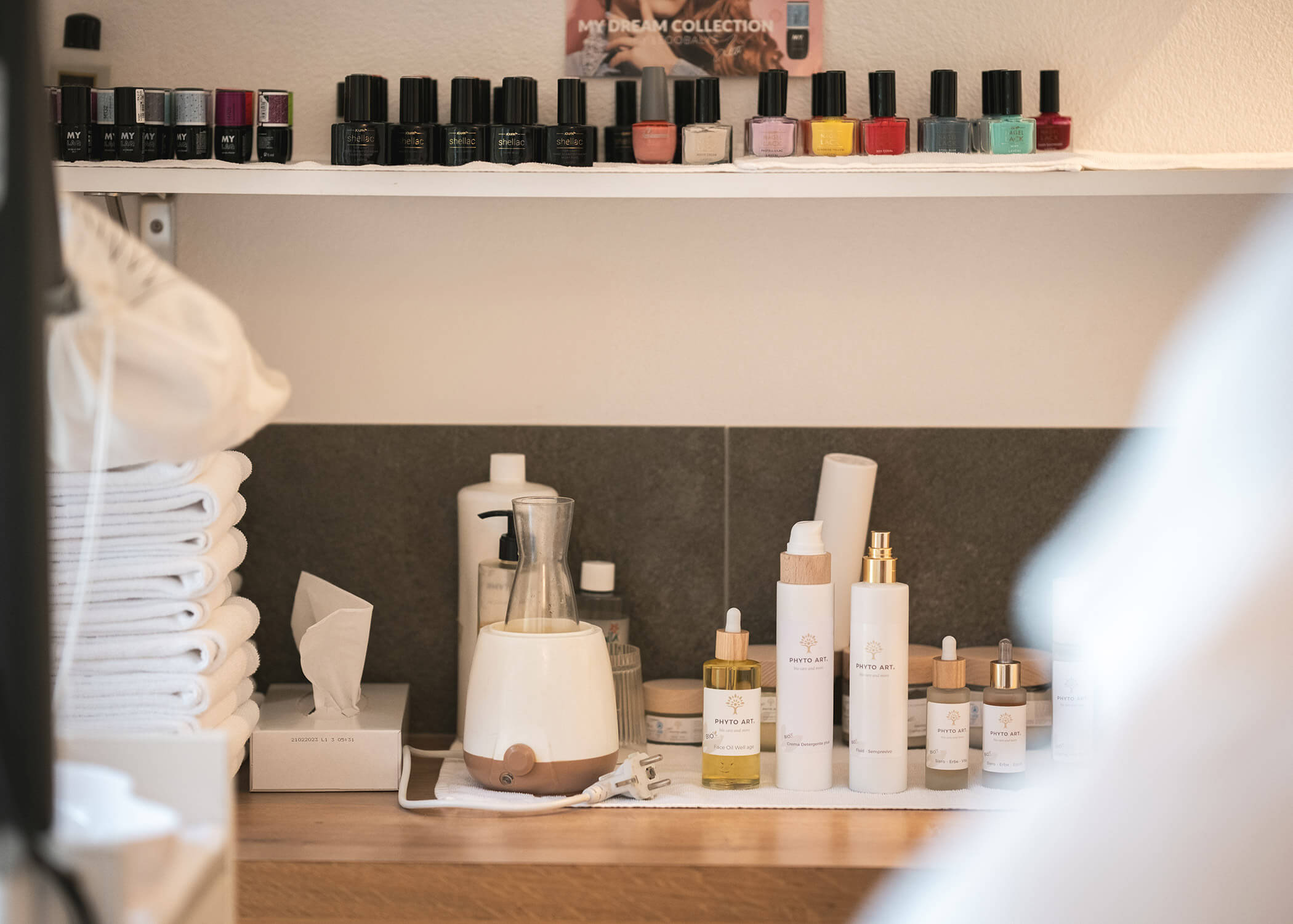 Ein Regal mit gefalteten Handtüchern, Flaschen, einem Diffusor und fein säuberlich angeordneten Nagellacken in verschiedenen Farben. - Hotel La Casies Mountain Living