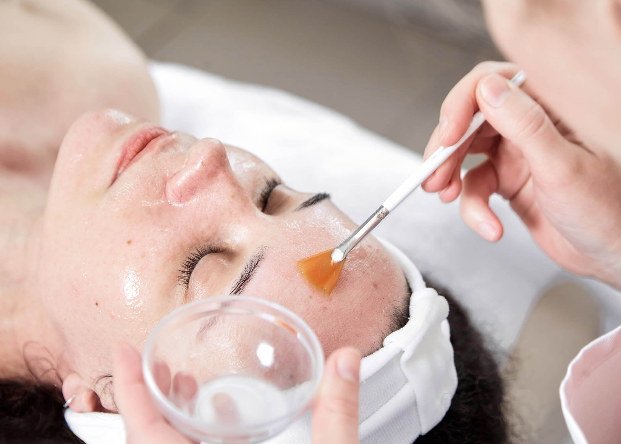 Eine Person erhält eine Gesichtsbehandlung mit einer Bürste, während sie liegt, die Augen geschlossen hat und ein weißes Stirnband trägt. - Hotel La Casies Mountain Living