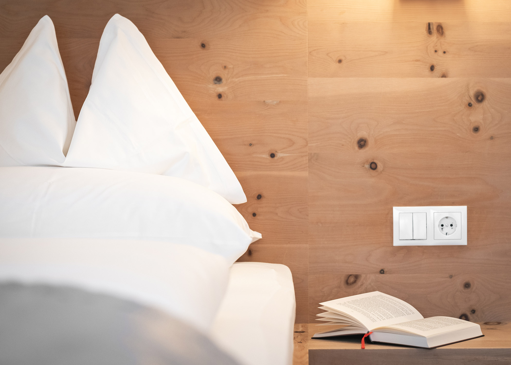 Ein Bett mit weißen Kissen und Bettwäsche, eine Holzwand und ein aufgeschlagenes Buch auf einem Nachttisch neben einer Steckdose. - Hotel La Casies Mountain Living