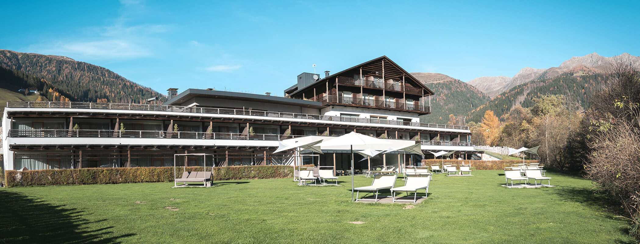 Modernes Berg-Resort mit Balkonen, Liegestühlen und Sonnenschirmen auf einer Wiese, umgeben von Bäumen und Hügeln. - Hotel La Casies Mountain Living