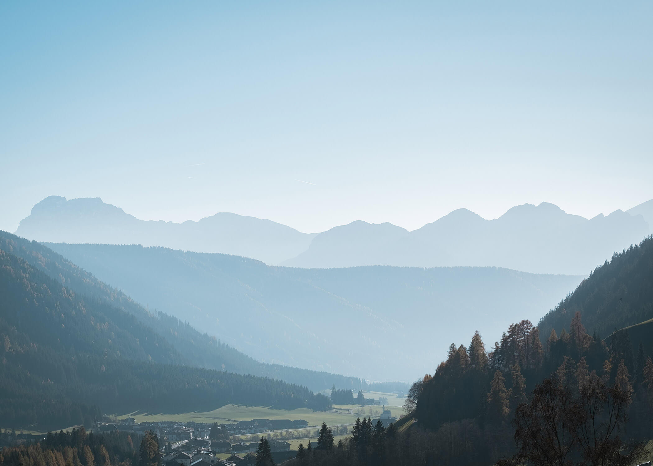 Berglandschaft mit bewaldeten Hügeln, entfernten Gipfeln und einem kleinen Dorf in einem grünen Tal unter klarem Himmel. - Hotel La Casies Mountain Living