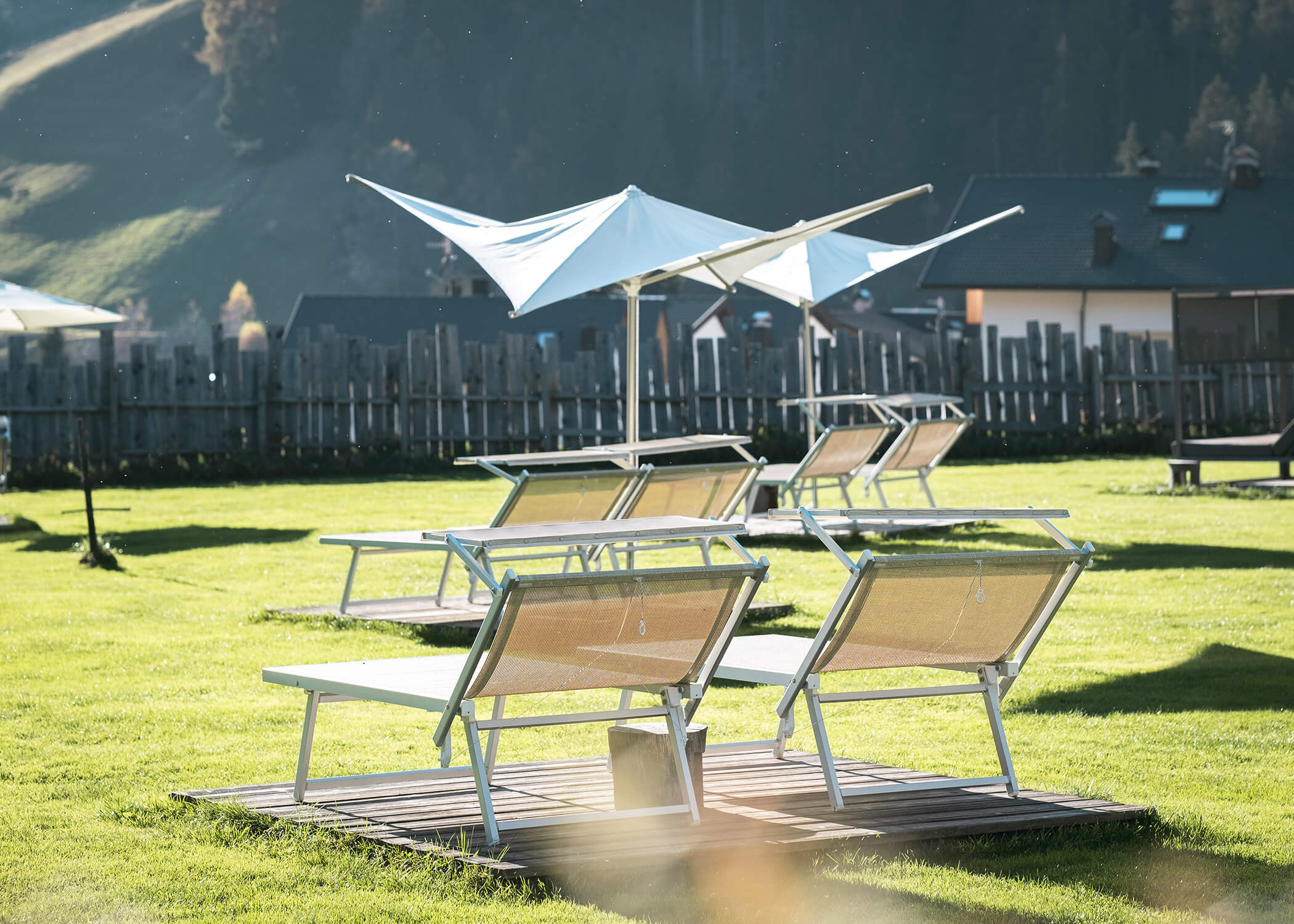 Liegestühle mit Sonnenschirmen auf einer Wiese, mit einem Holzzaun und Gebäuden im Hintergrund. - Hotel La Casies Mountain Living