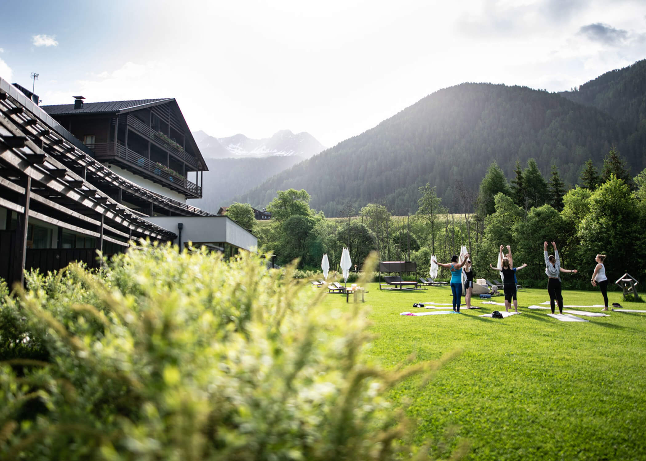 Eine Gruppe von Menschen macht Yoga auf einer Wiese in der Nähe eines Gebäudes, umgeben von Bergen und Bäumen. - Hotel La Casies Mountain Living