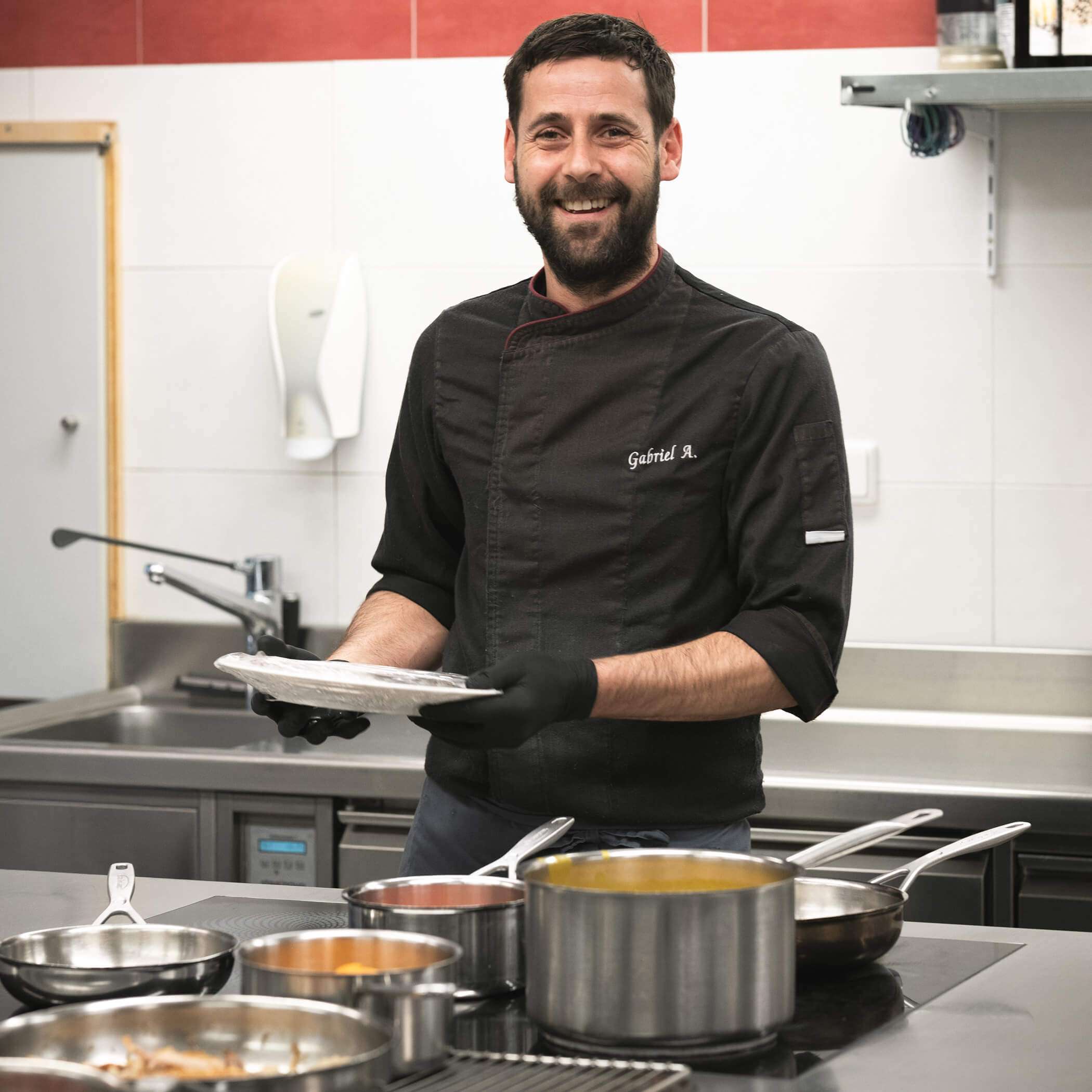 Ein lächelnder Koch in schwarzer Uniform steht in einer Küche und hält einen Teller neben Töpfen und Pfannen auf dem Herd. - Hotel La Casies Mountain Living