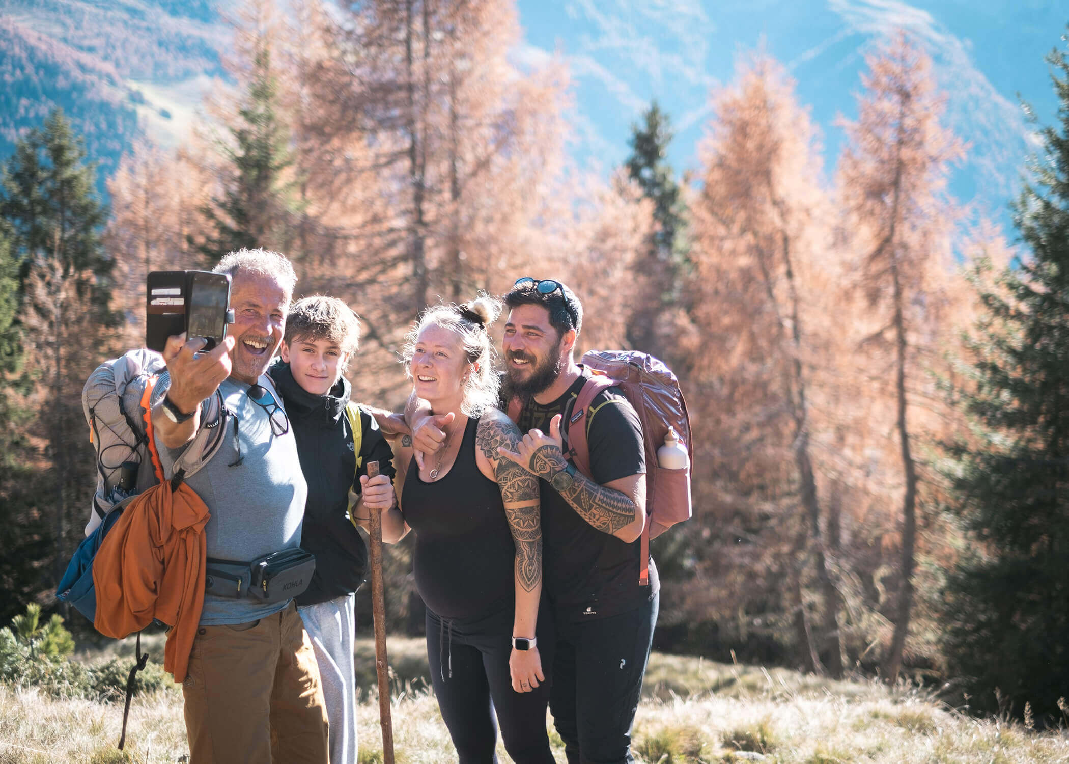 Vier Personen mit Rucksäcken lächeln und machen ein Selfie, während sie in einer sonnenbeschienenen, bewaldeten Bergregion wandern. - Hotel La Casies Mountain Living