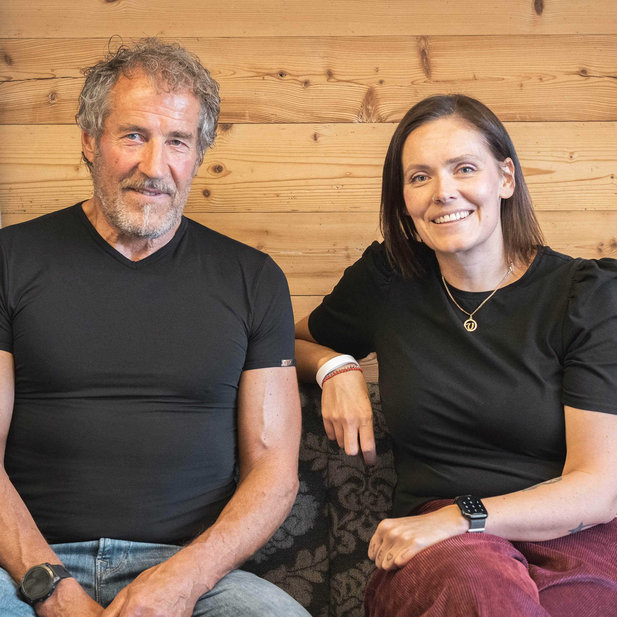 Zwei Personen sitzen lächelnd auf einer Couch vor einer Holzwand. - Hotel La Casies Mountain Living