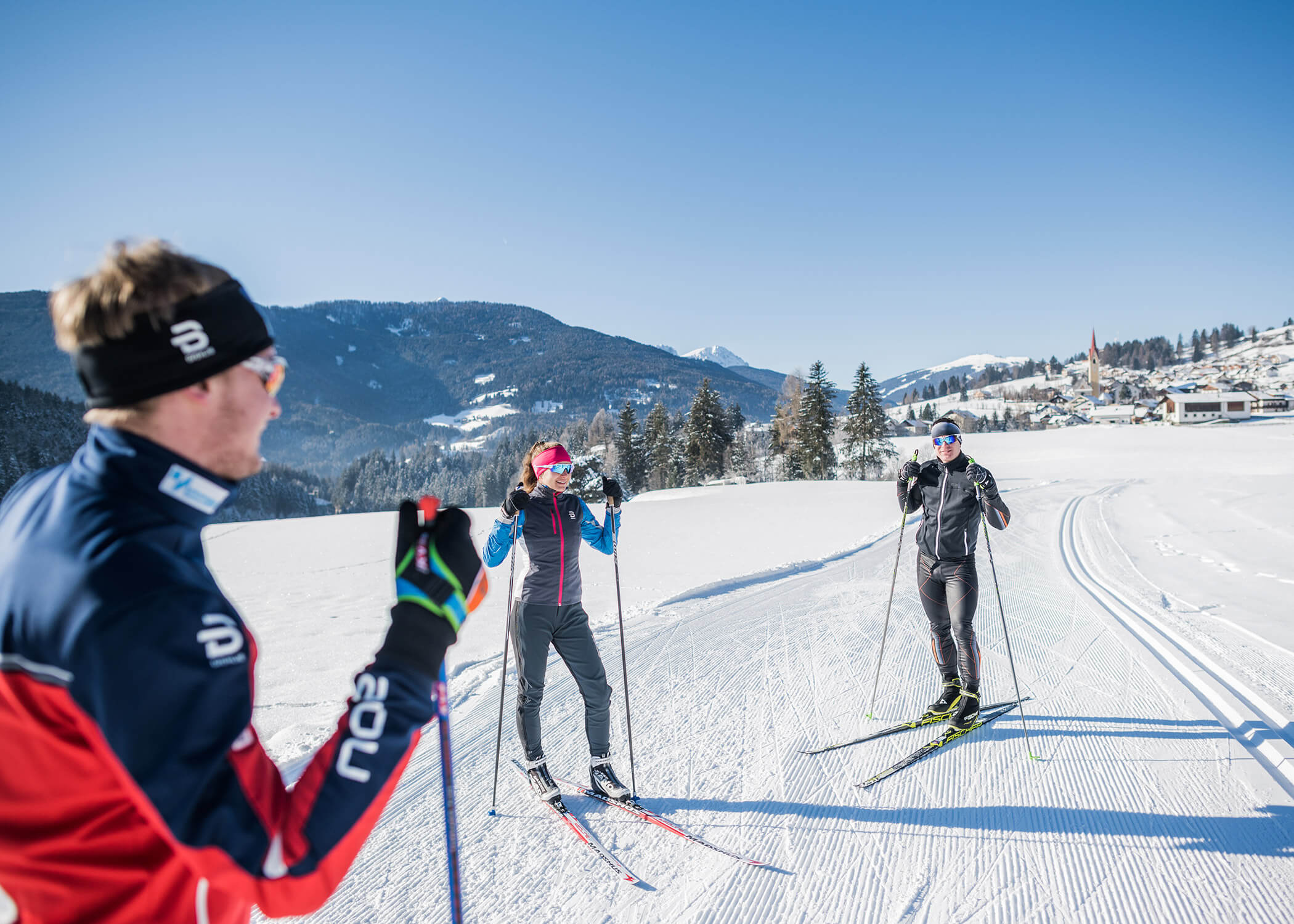 Drei Personen beim Skilanglauf in einer sonnigen, verschneiten Landschaft mit Bergen und einem Dorf im Hintergrund. - Hotel La Casies Mountain Living