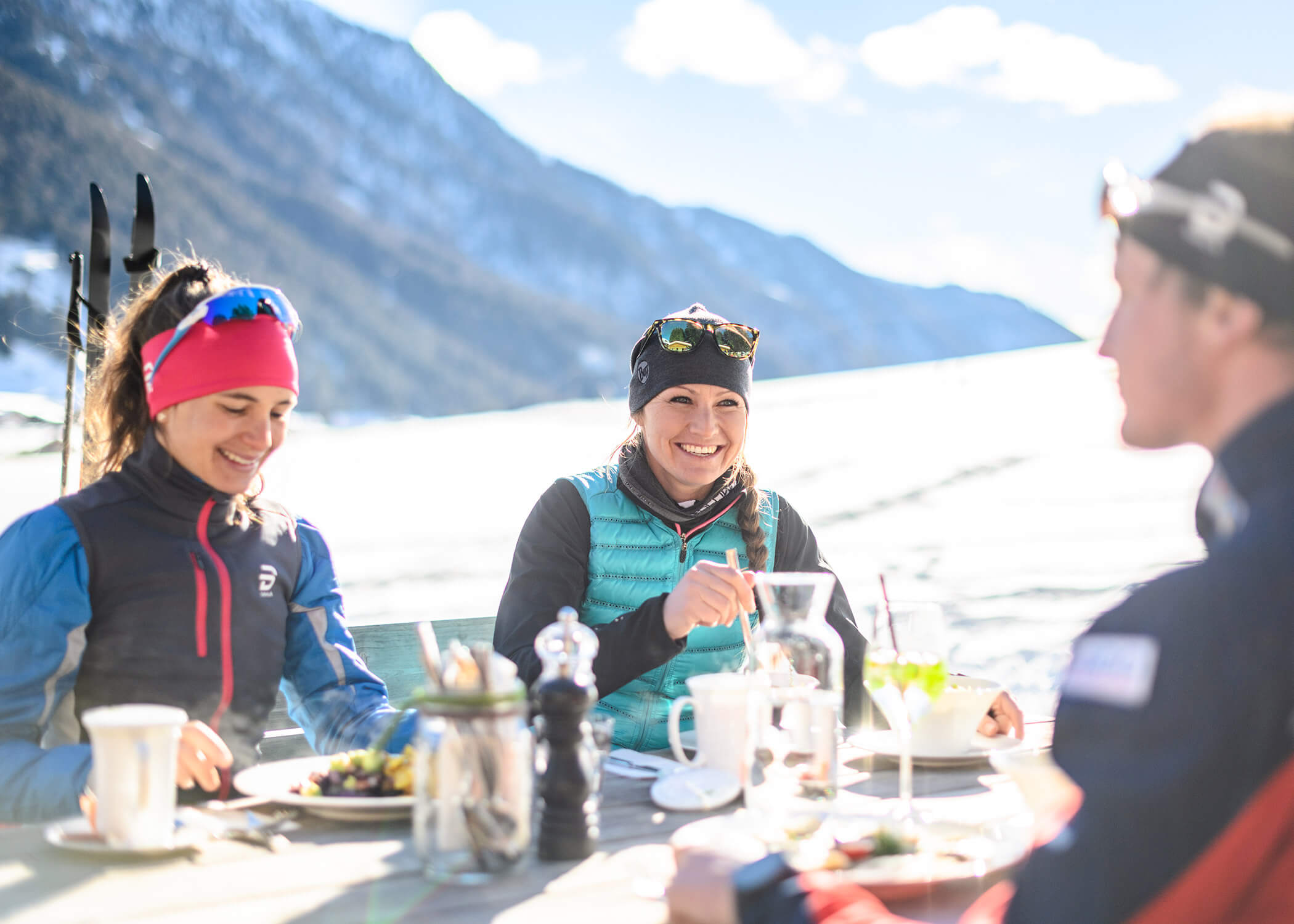 Drei Personen in Wintersportkleidung essen und lächeln an einem Tisch im Freien mit verschneiten Bergen im Hintergrund. - Hotel La Casies Mountain Living