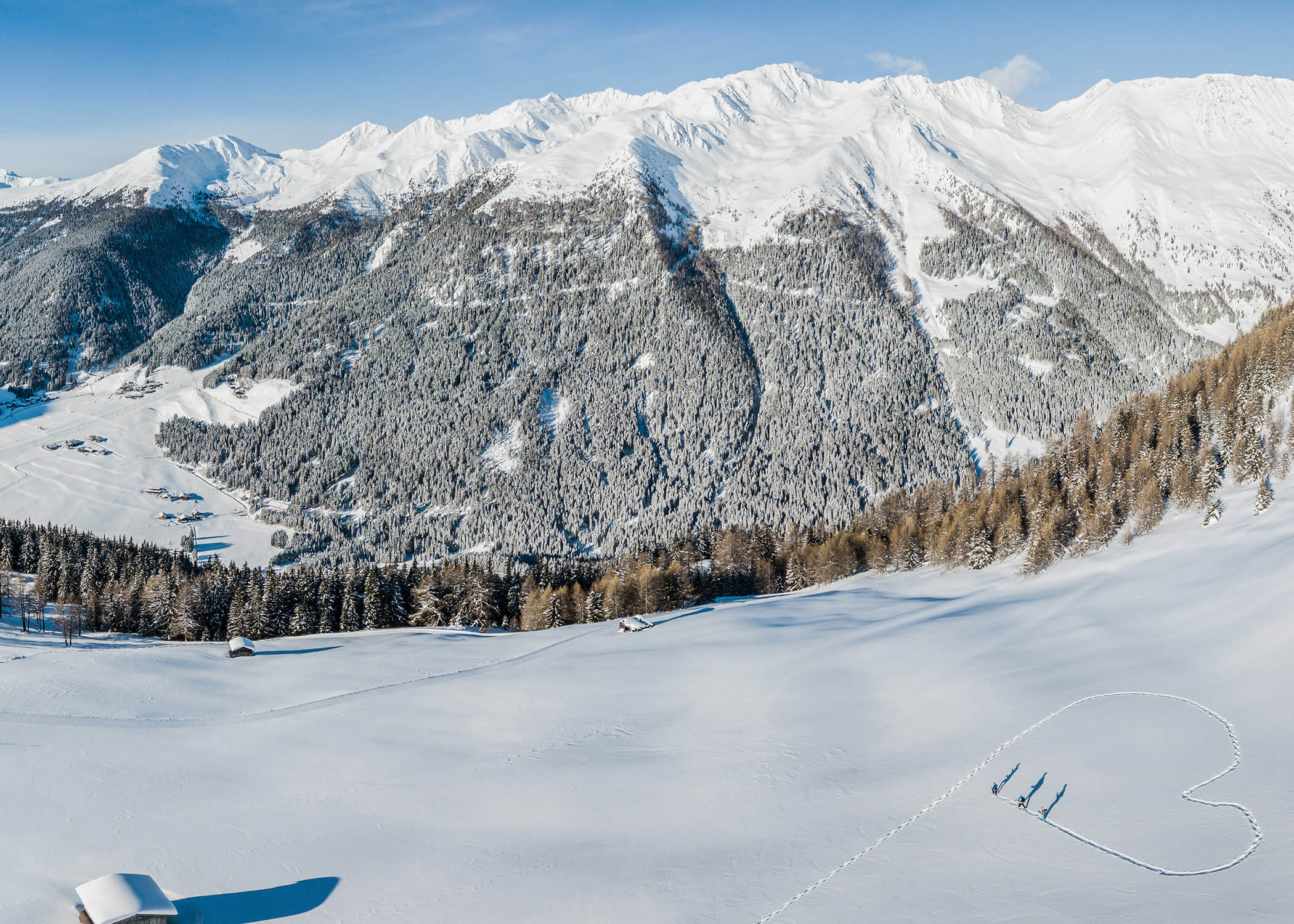 Eine verschneite Berglandschaft mit einer großen, in den Schnee gezeichneten Herzform und drei in der Nähe stehenden Personen. - Hotel La Casies Mountain Living