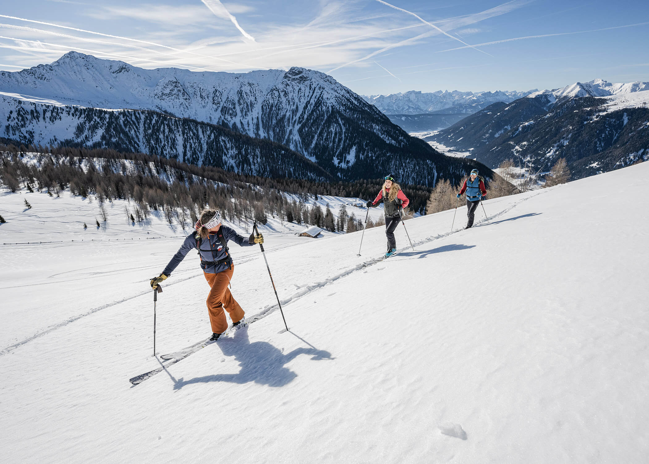 Drei Personen fahren auf einem verschneiten Berghang mit fernen Bergen unter einem klaren Himmel bergauf. - Hotel La Casies Mountain Living