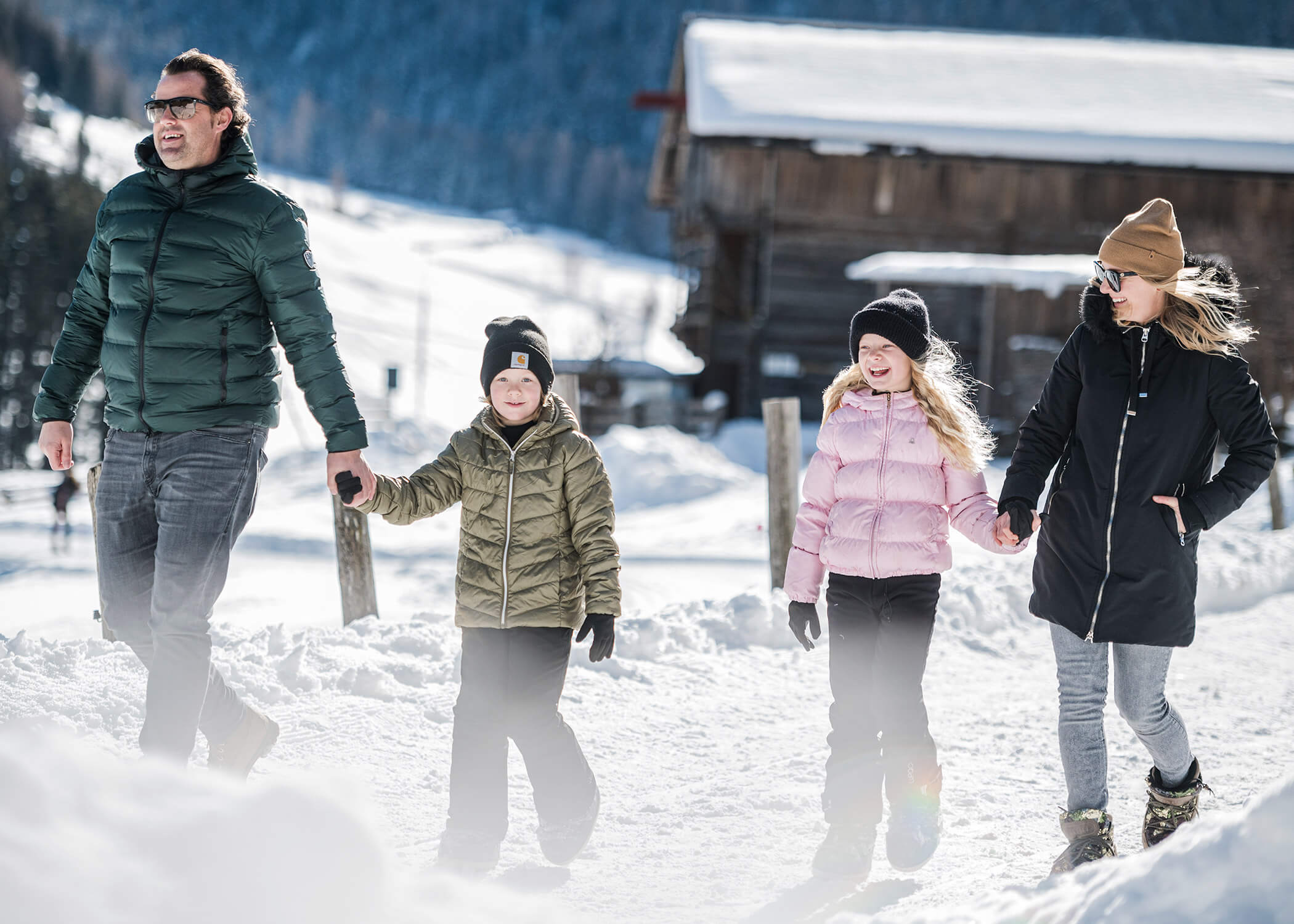 Eine vierköpfige Familie spaziert in Wintermänteln und Mützen durch eine verschneite Landschaft, hält sich an den Händen und lächelt. - Hotel La Casies Mountain Living