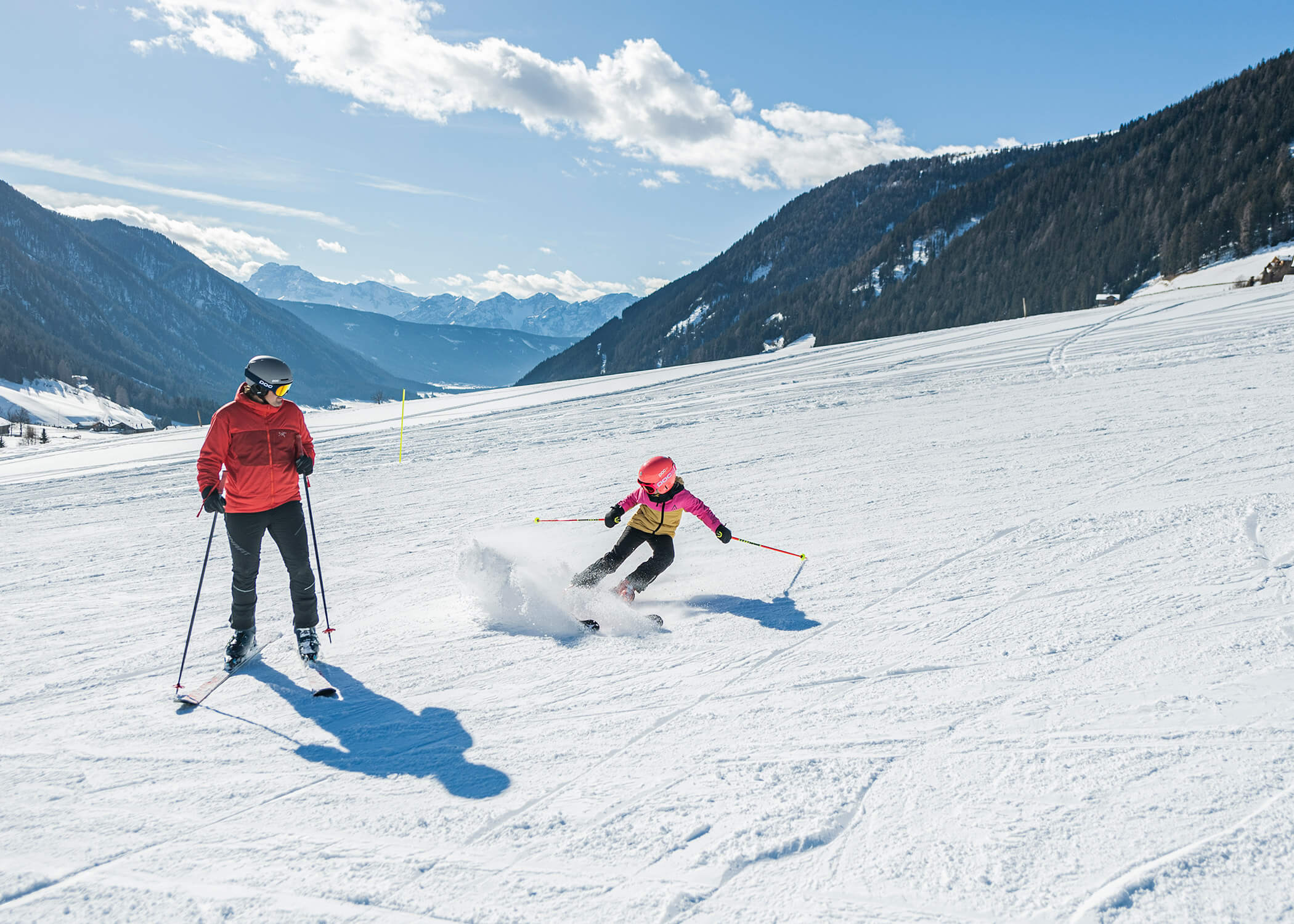 Zwei Personen beim Skifahren auf einem verschneiten Berghang unter einem klaren blauen Himmel mit Bergen im Hintergrund. - Hotel La Casies Mountain Living