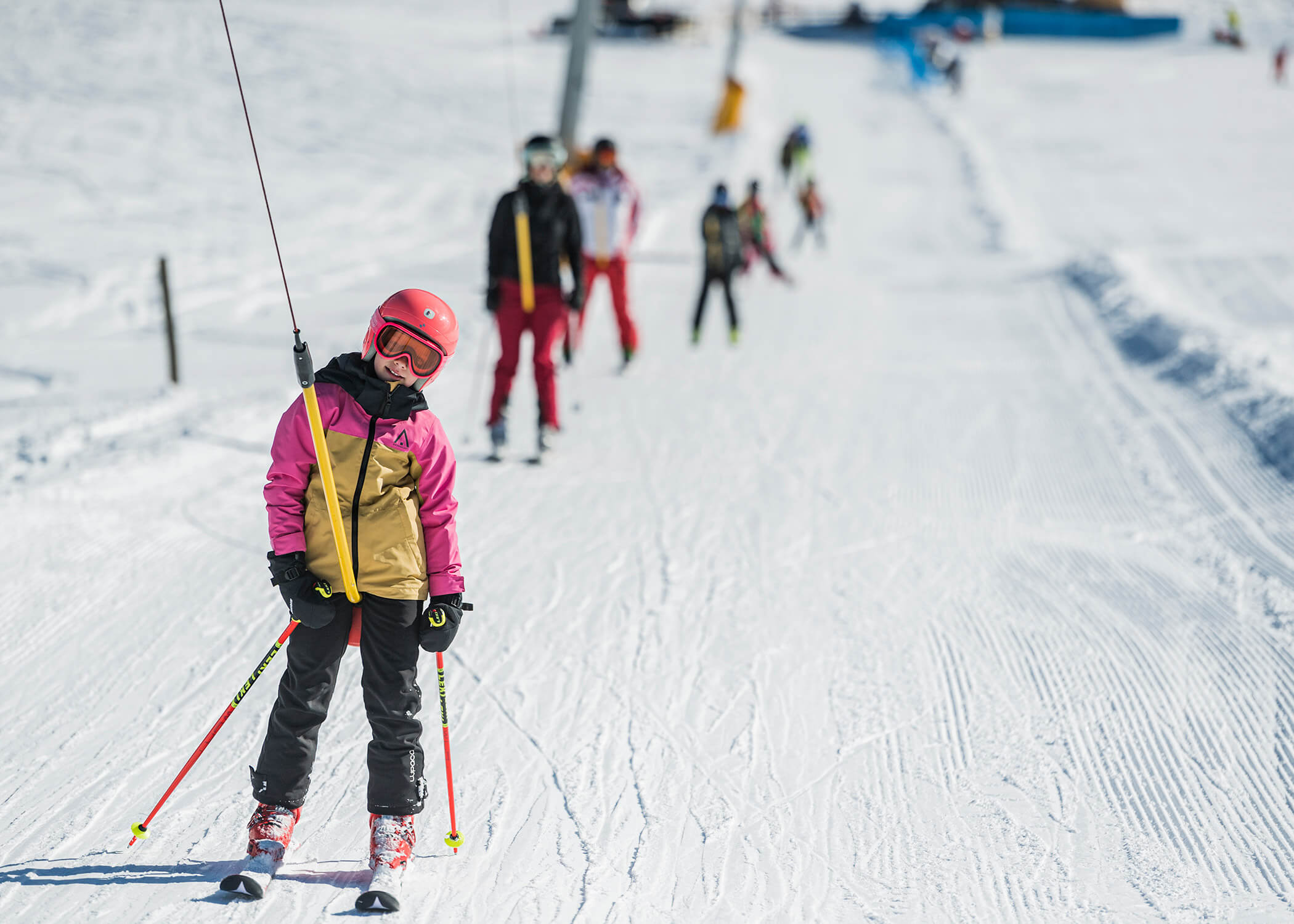Kind in rosafarbener Skiausrüstung, das einen Skilift benutzt, mit anderen auf der verschneiten Piste im Hintergrund. - Hotel La Casies Mountain Living