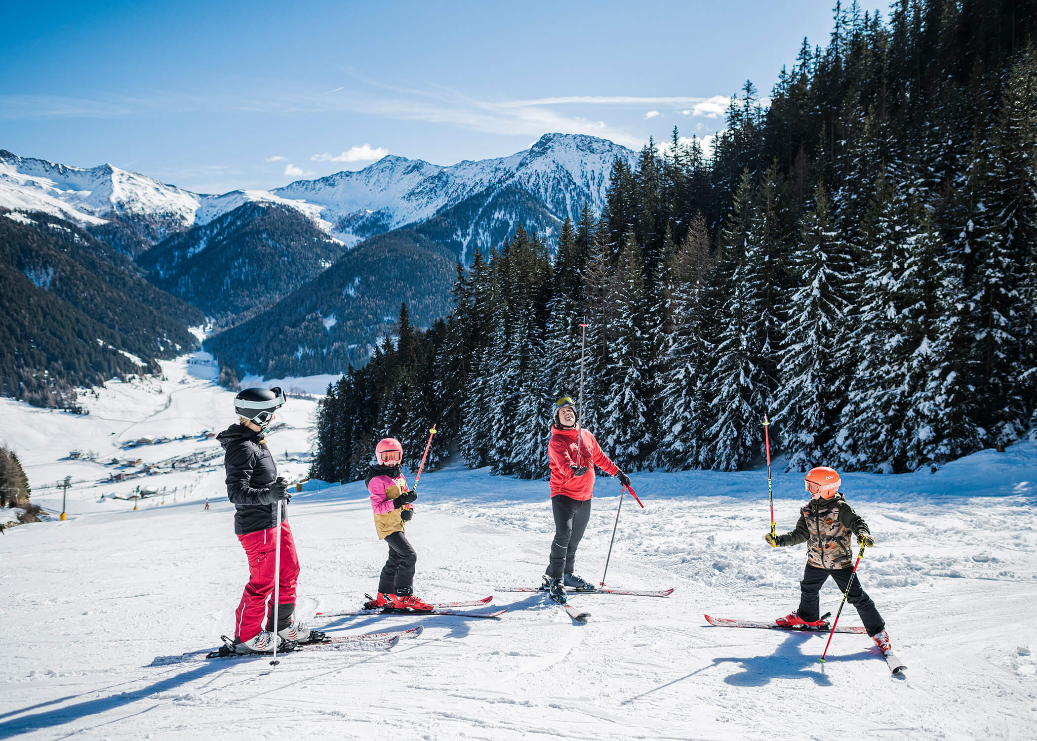Vier Personen beim Skifahren auf einem verschneiten Berghang mit Kiefern und schneebedeckten Gipfeln im Hintergrund. - Hotel La Casies Mountain Living