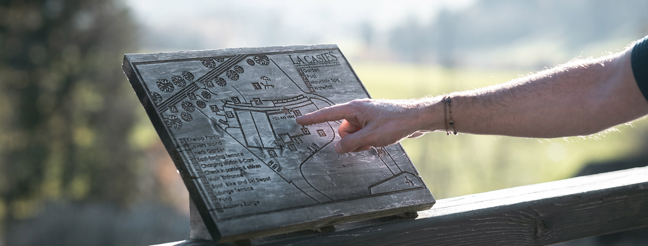 Eine Person zeigt auf eine Holzkarte im Freien mit unscharfer Landschaft im Hintergrund. - Hotel La Casies Mountain Living