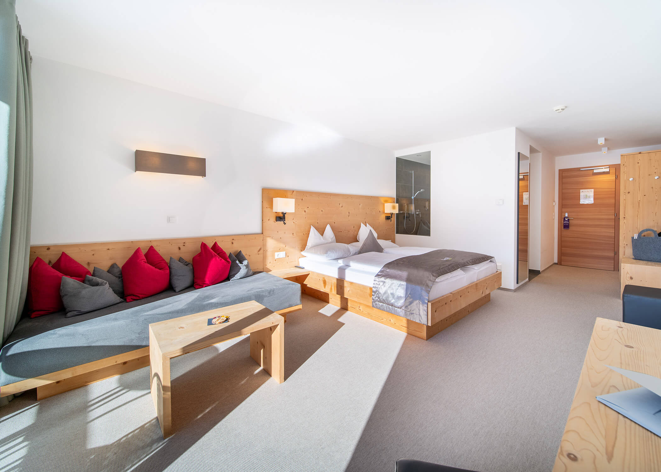 Helles Hotelzimmer mit einem Doppelbett, einem Sofa, einem Tisch, Holzakzenten und einem großen Fenster mit Tageslicht. - Hotel La Casies Mountain Living